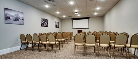 A conference room with rows of beige chairs, a carpeted floor, a projector screen, and artwork on the walls.