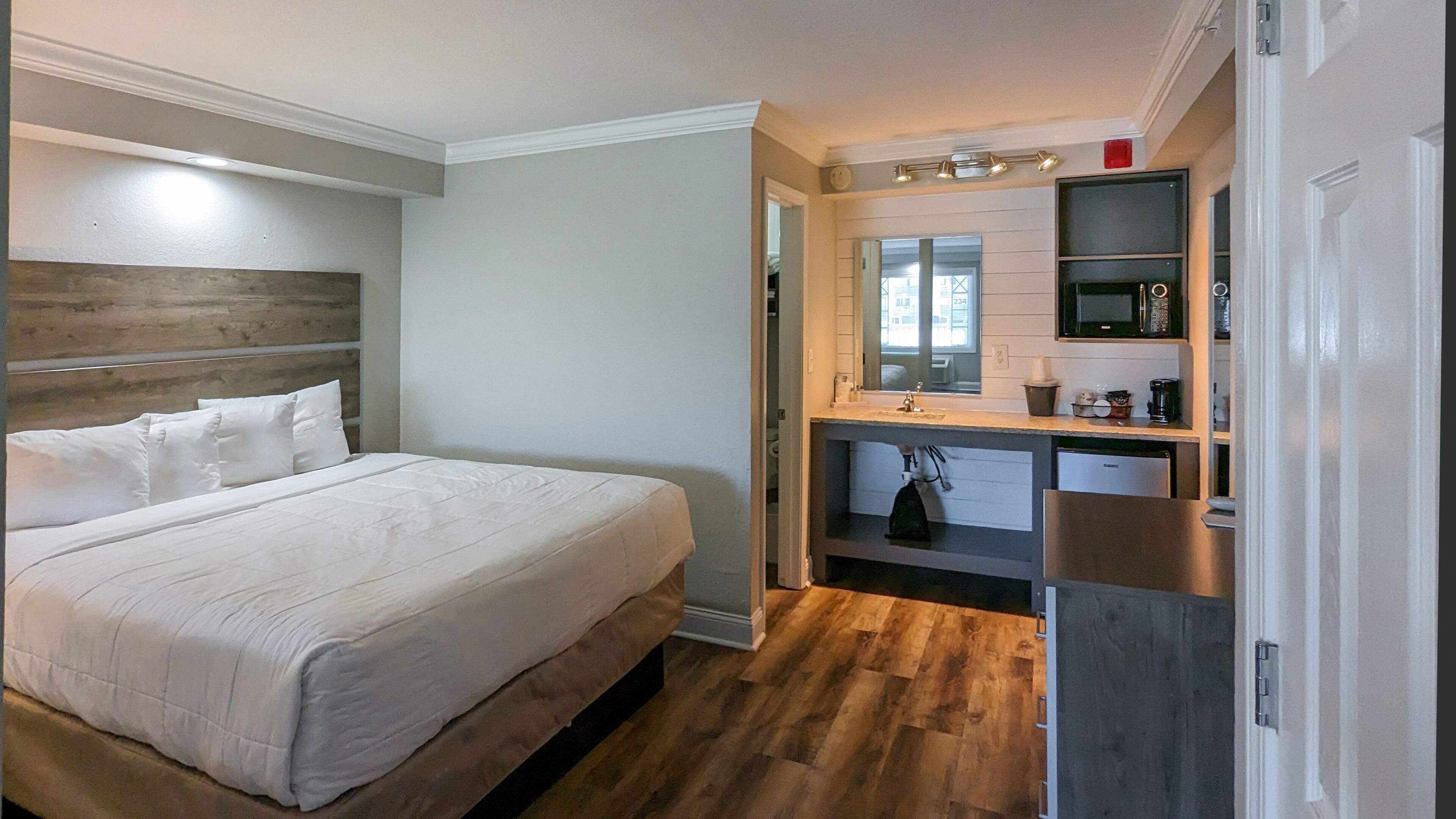 A bedroom with a large bed, a wooden headboard, a built-in desk with a mirror, and a kitchenette area.