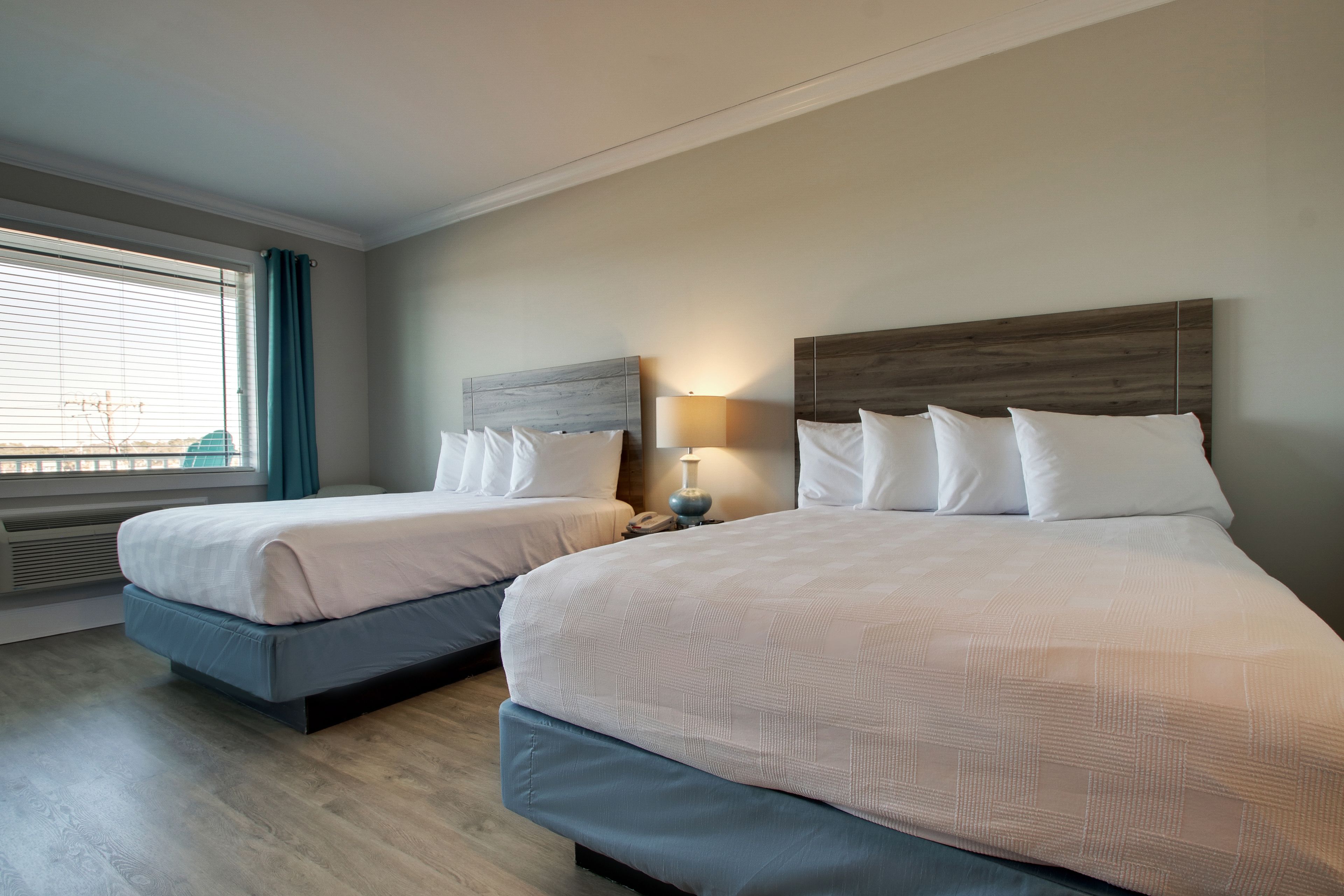 A hotel room with two beds, a window with curtains, a bedside table with a lamp, and a wall-mounted heater.