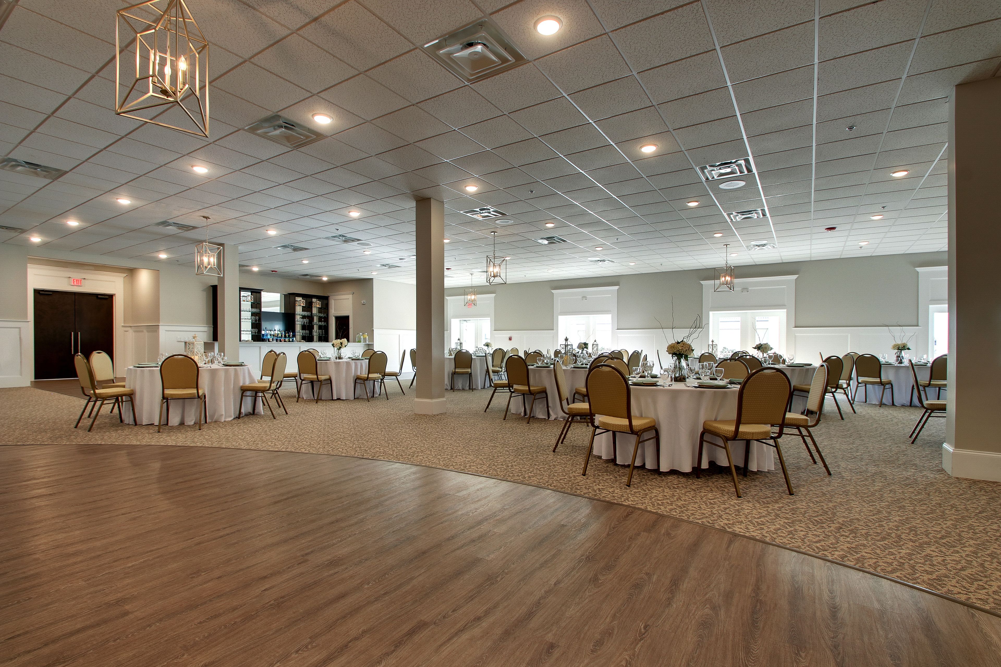 A spacious banquet hall with multiple round tables and chairs, a high ceiling with recessed lighting, and a carpeted floor.