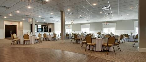 A spacious banquet hall with multiple round tables and chairs, a high ceiling with recessed lighting, and a carpeted floor.