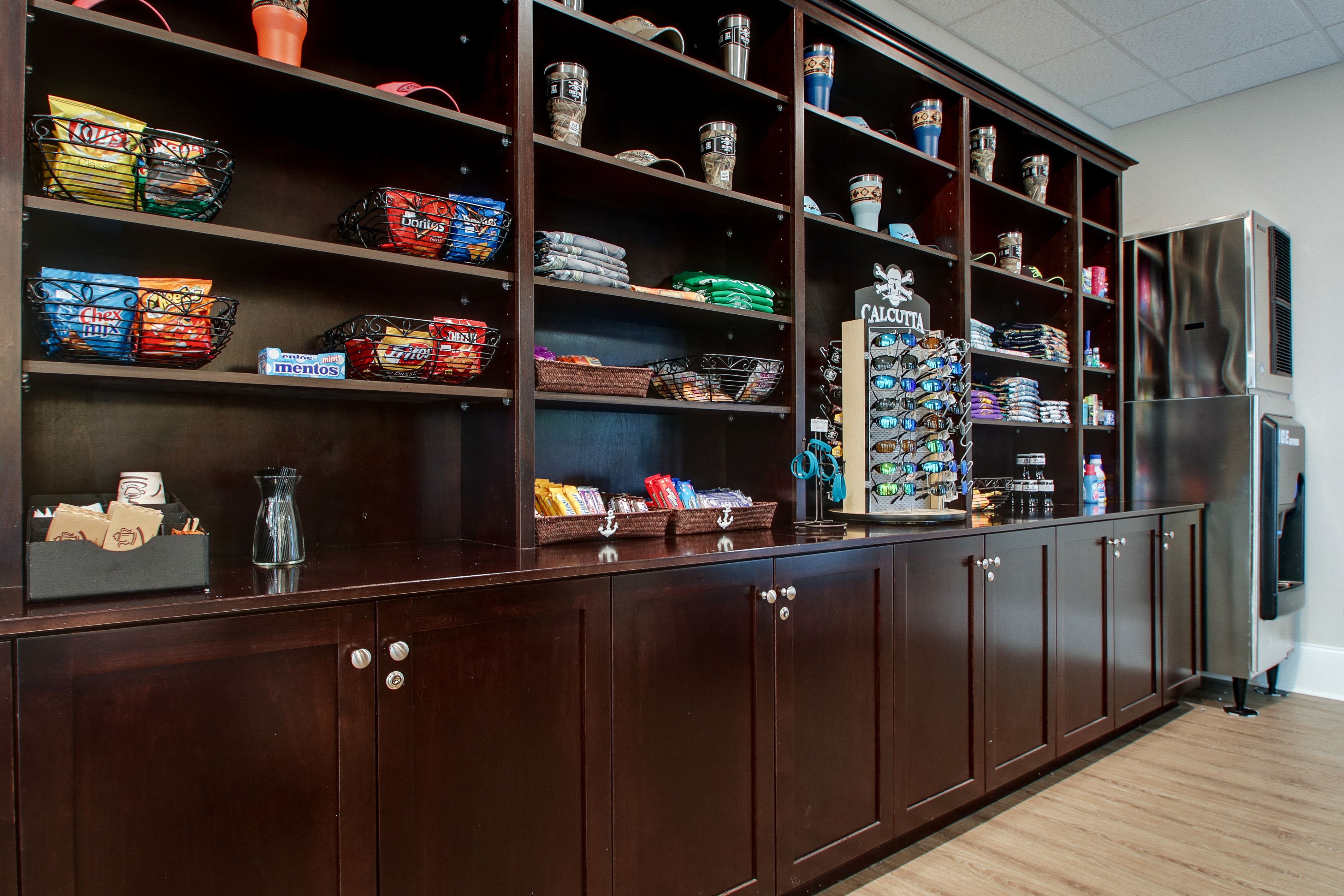 A kitchen with wooden cabinets, a stainless steel refrigerator, and shelves stocked with snacks and kitchenware.