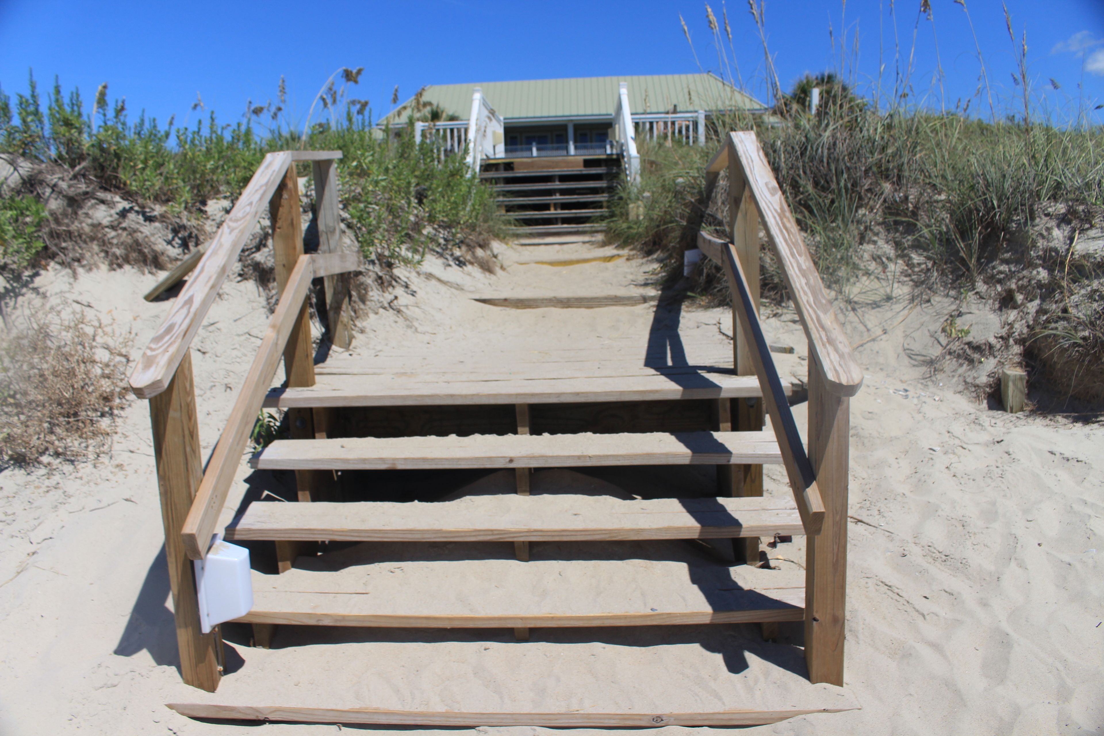 A wooden staircase leading to a building with a green roof, surrounded by sand and sparse vegetation.