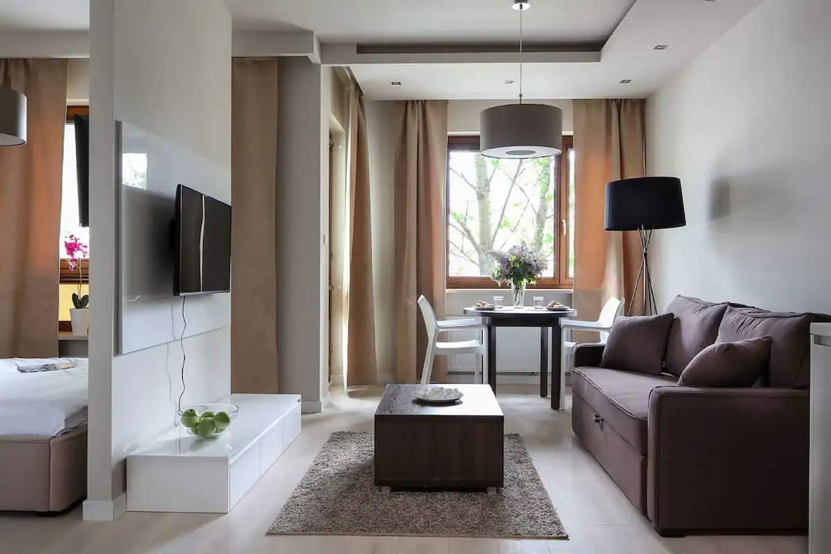 Una habitación de hotel moderna con sofá, televisión instalada en la pared, mesa de comedor con sillas y un amplio ventanal con cortinas.