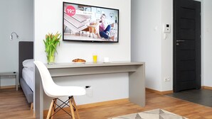 Et moderne hotelværelse med et gråt skrivebord, en hvid stol, et fladskærms-tv monteret på væggen, en lille plante og et morgenmadssæt med croissanter og juice.