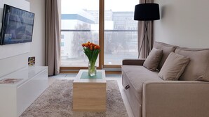 A modern living room with a large window, a flat-screen TV mounted on the wall, a beige sofa, a glass coffee table, and a vase with flowers. - Chopin Apartments City (Warsaw)