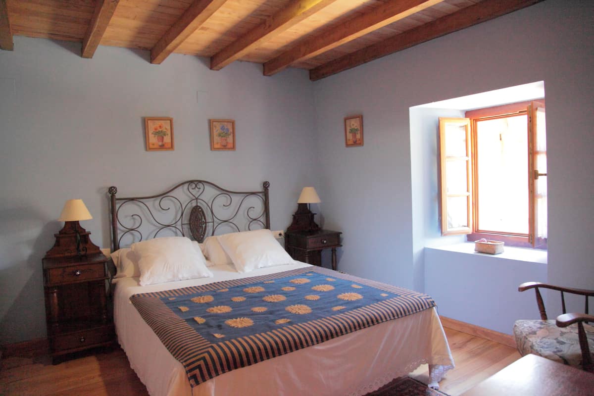 A bedroom with a wooden ceiling, a bed with a floral bedspread, two bedside tables with lamps, and a window with a view.