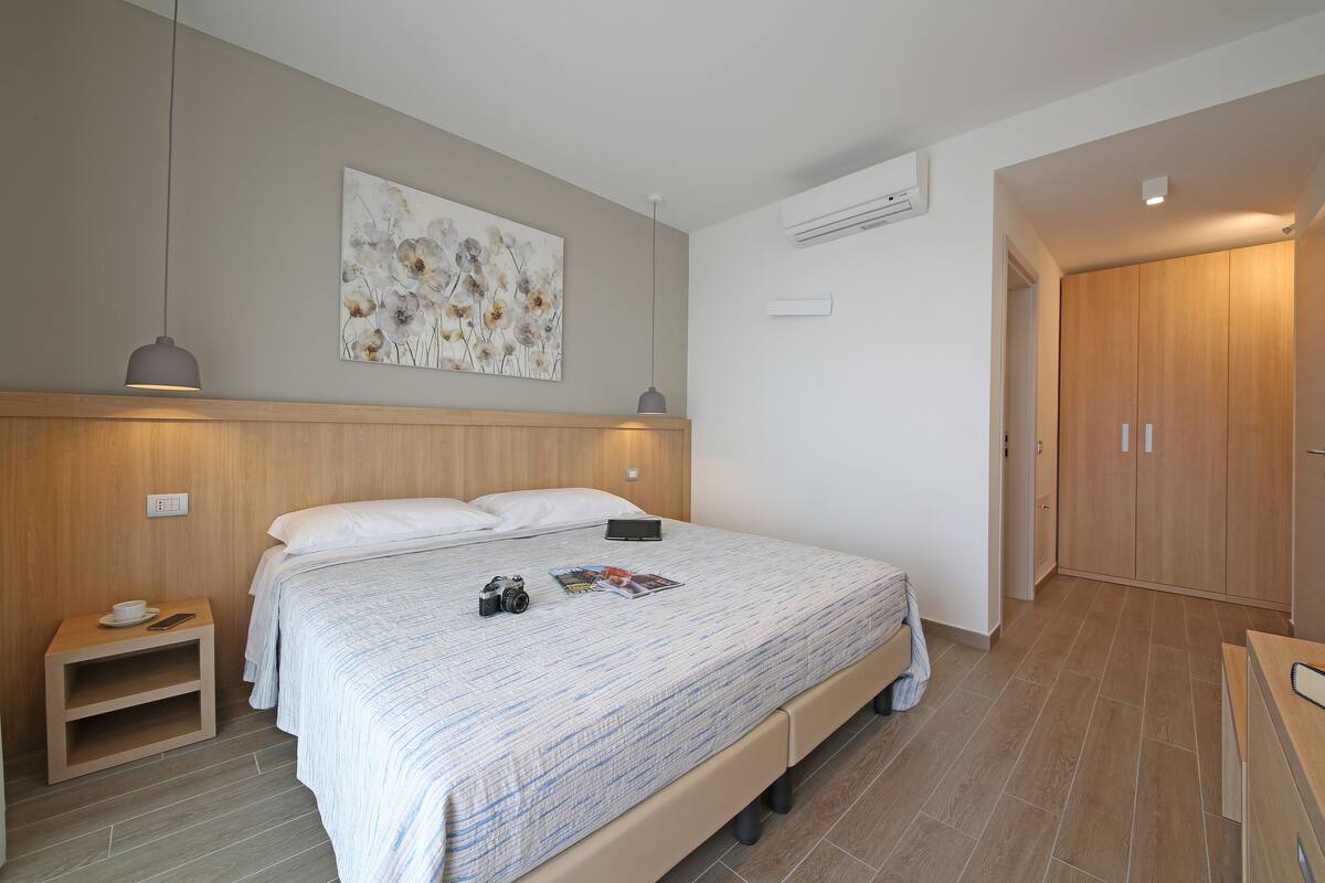 A bedroom with a wooden headboard, a bed with a patterned bedspread, bedside tables with a cup and a camera, and a painting on the wall.