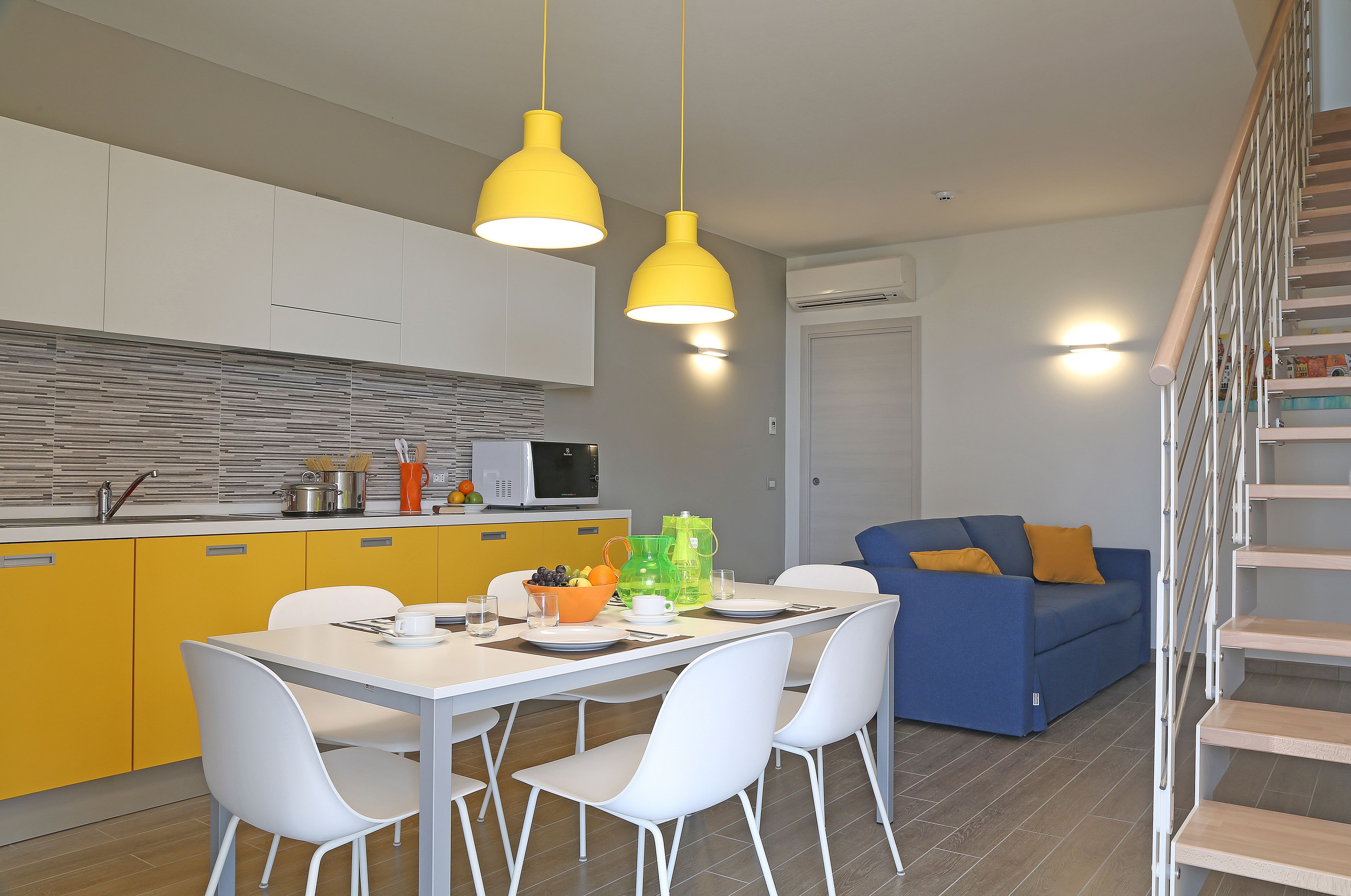 Une cuisine moderne avec des meubles de couleur jaune, une table à manger avec des chaises blanches et un canapé bleu dans l’espace salon.
