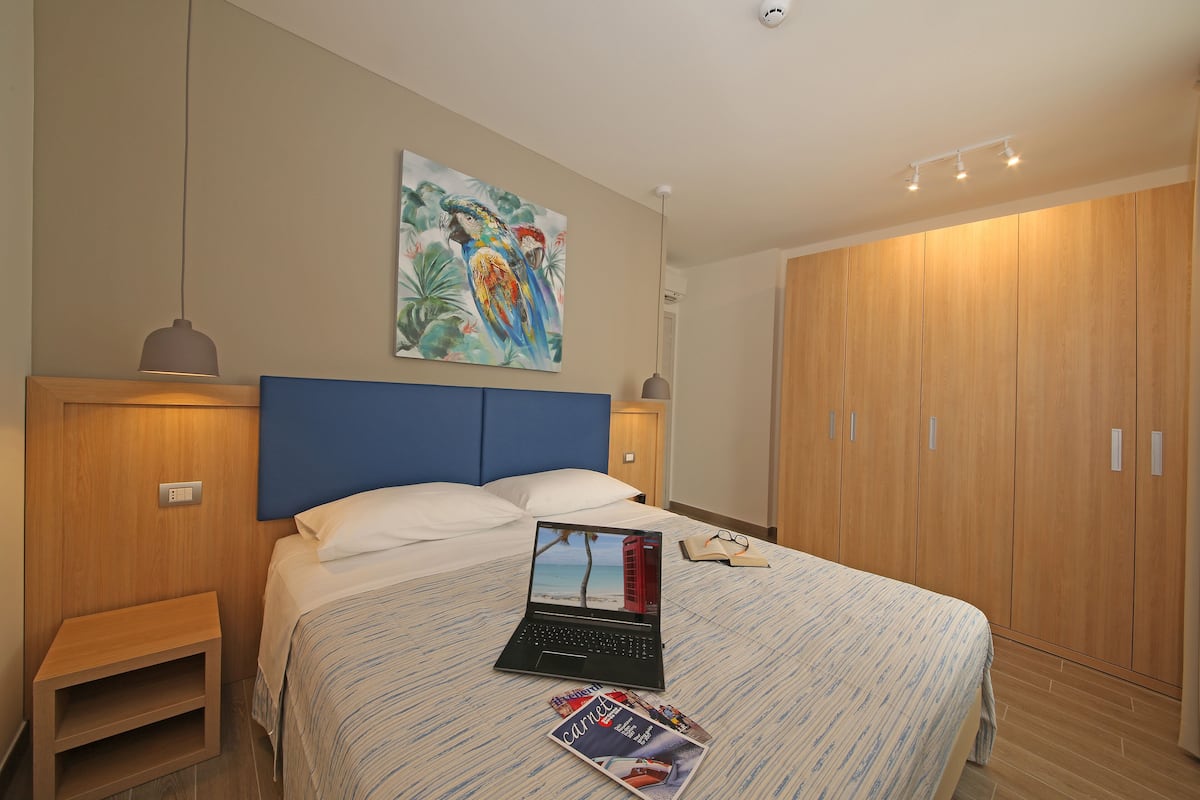 A bedroom with a bed, a laptop, a magazine, and a painting on the wall.