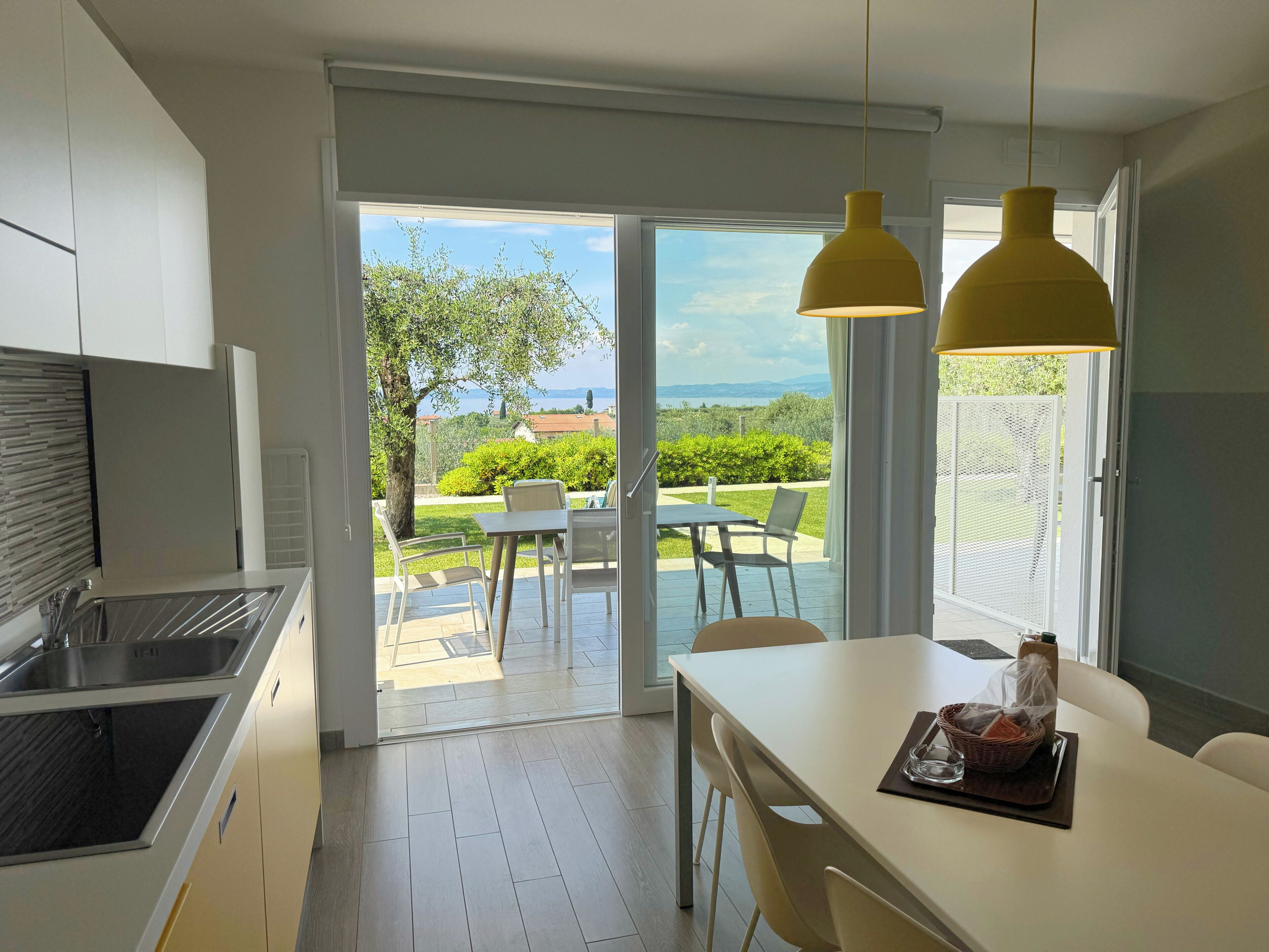 Une cuisine moderne dotée d’un four encastré, un coin repas avec une table et des chaises, et une vue sur un jardin et des montagnes grâce à de grandes portes vitrées.