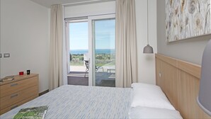 Een hotelkamer met een bed, een houten dressoir, een bureau met stoel, en uitzicht op de omgeving via een schuifdeur.