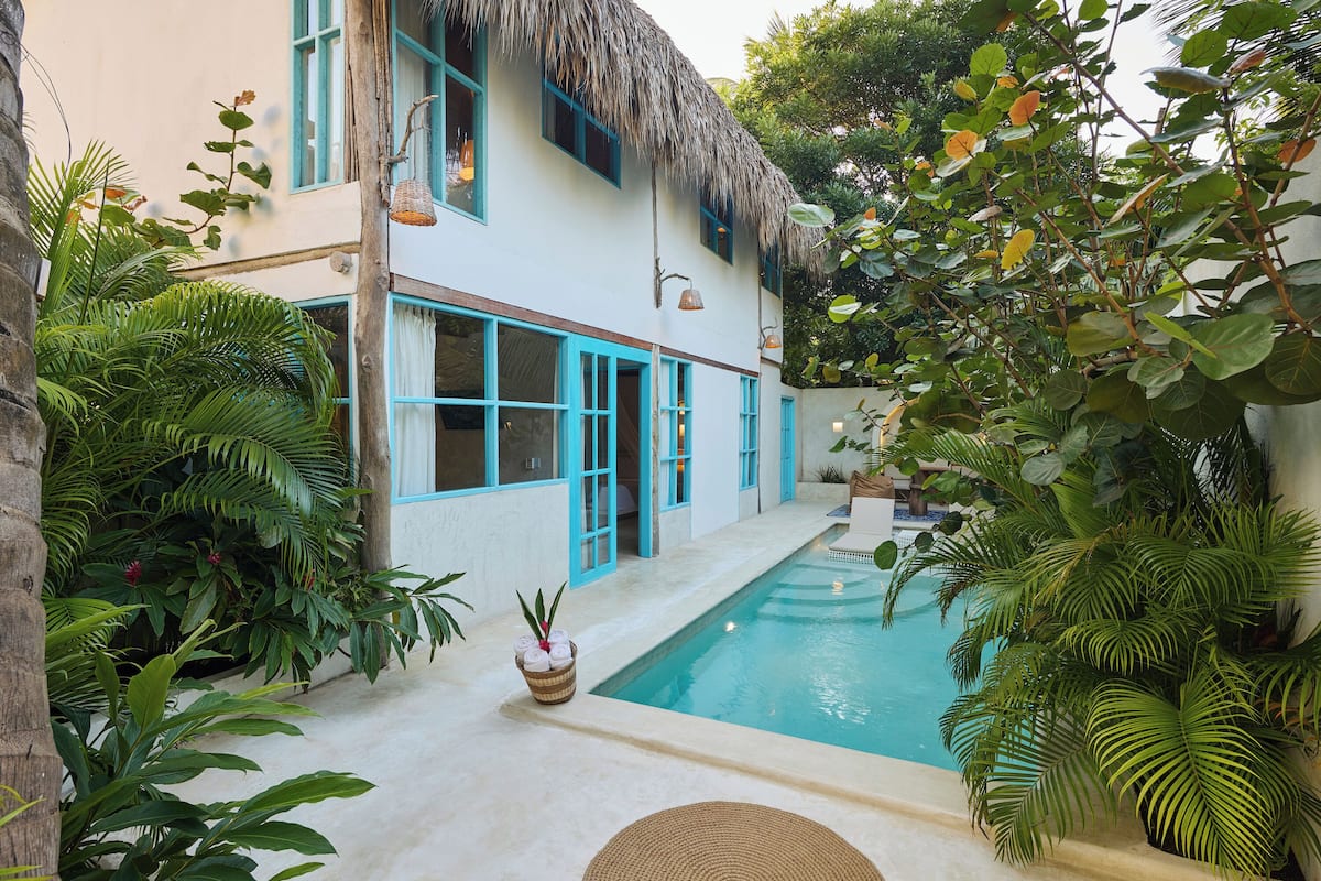 Vue sur un bâtiment à toit de chaume, pris depuis la piscine, et entouré d’une végétation tropicale luxuriante.
