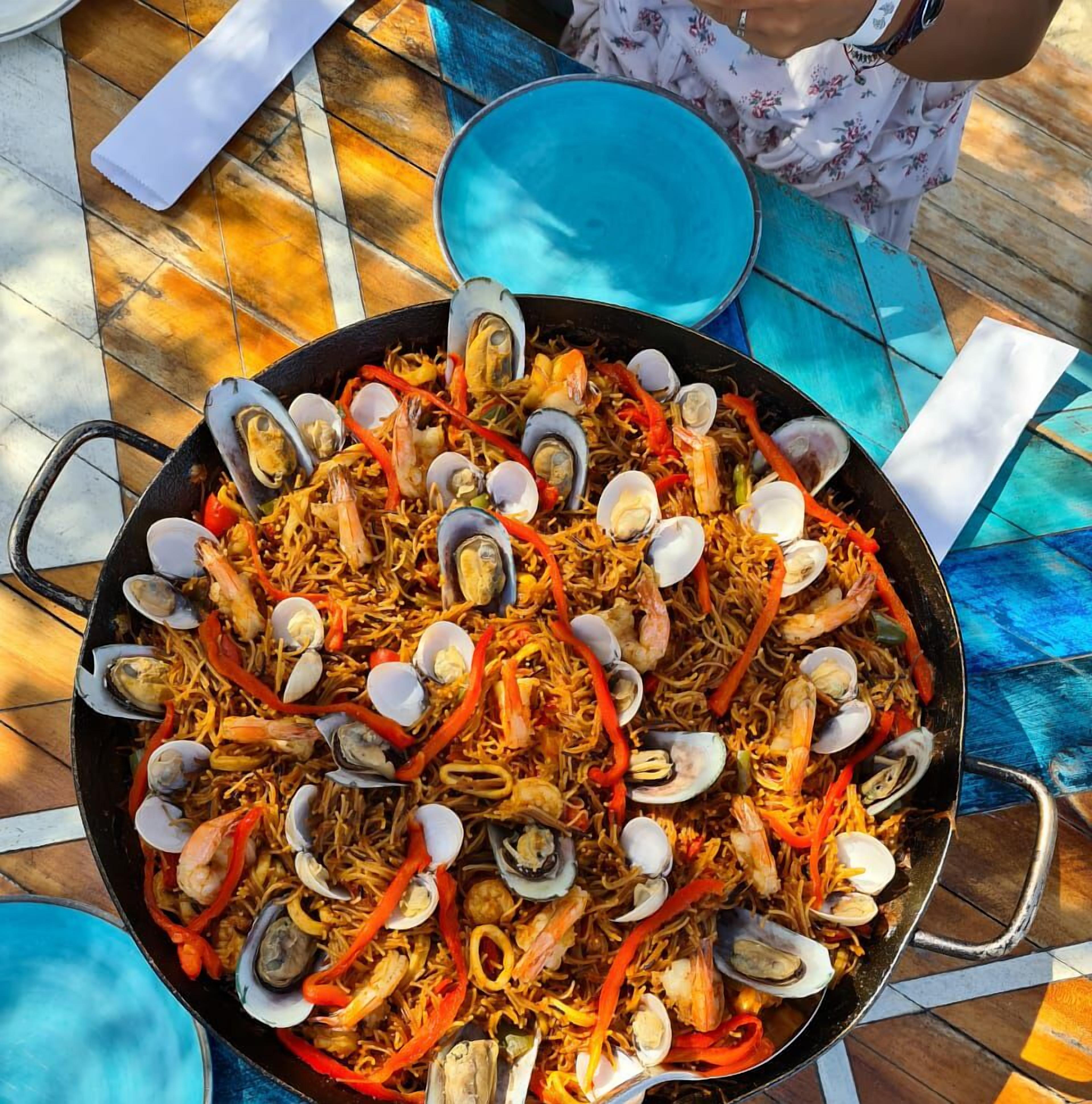 A large seafood paella dish on a table with a tiled floor and a blue chair.