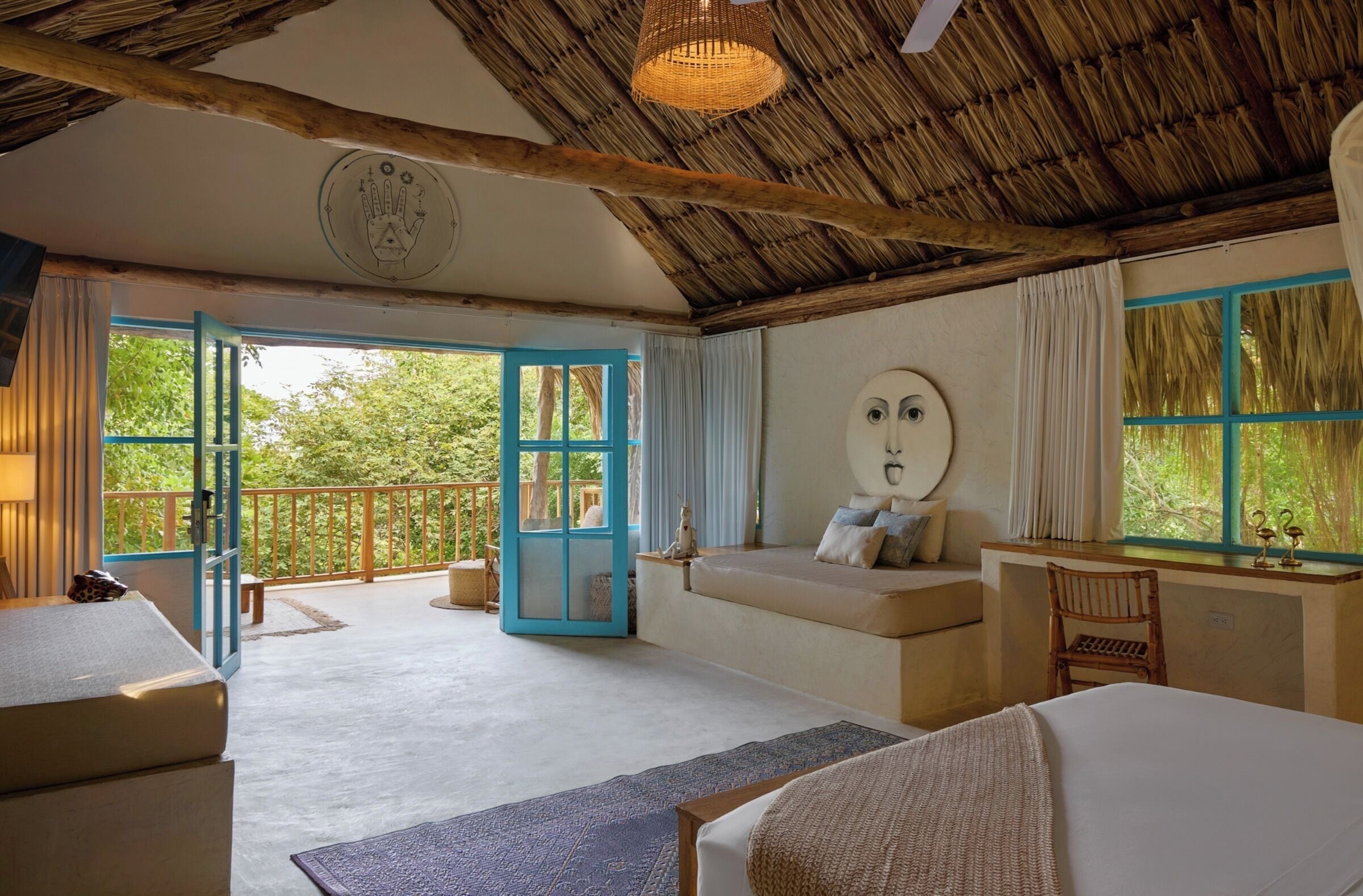 A room with a thatched roof, a large window with blue shutters, a sofa, a bed, and a desk with a chair.