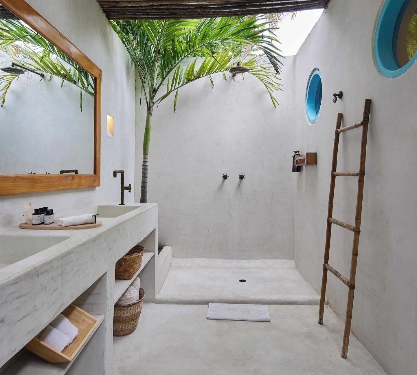 A modern bathroom with a large mirror, a wooden ladder, a sink with toiletries, and a shower area.