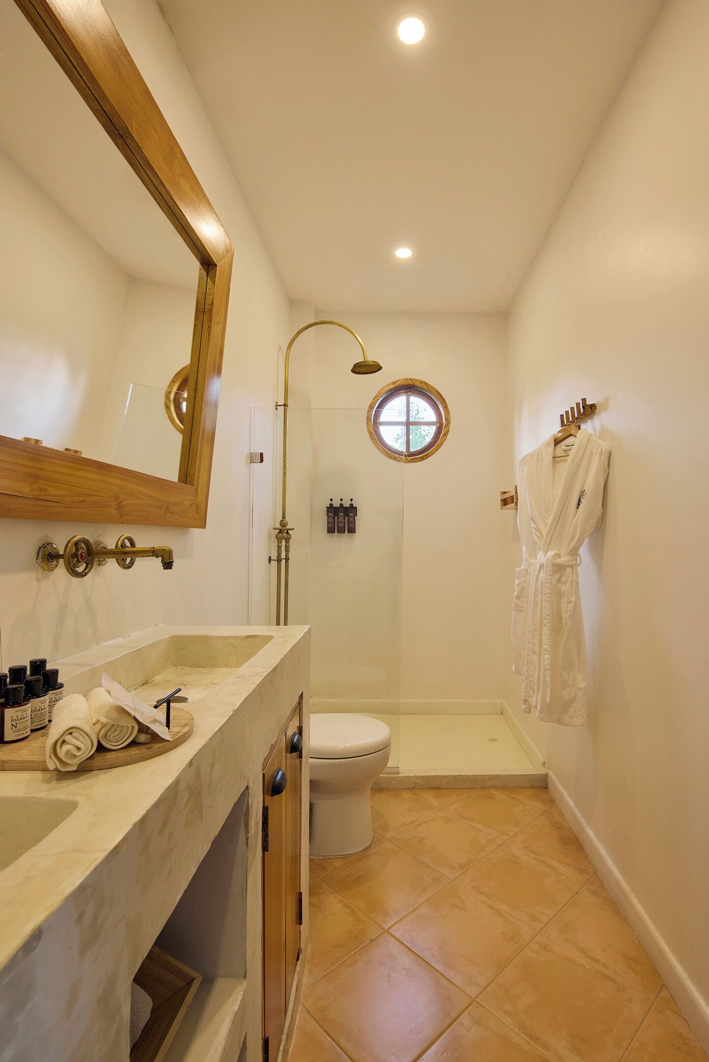 Une salle de bain avec un lavabo en marbre, un miroir rond, des toilettes blanches, une douche avec une porte en verre et une robe blanche accrochée au mur.