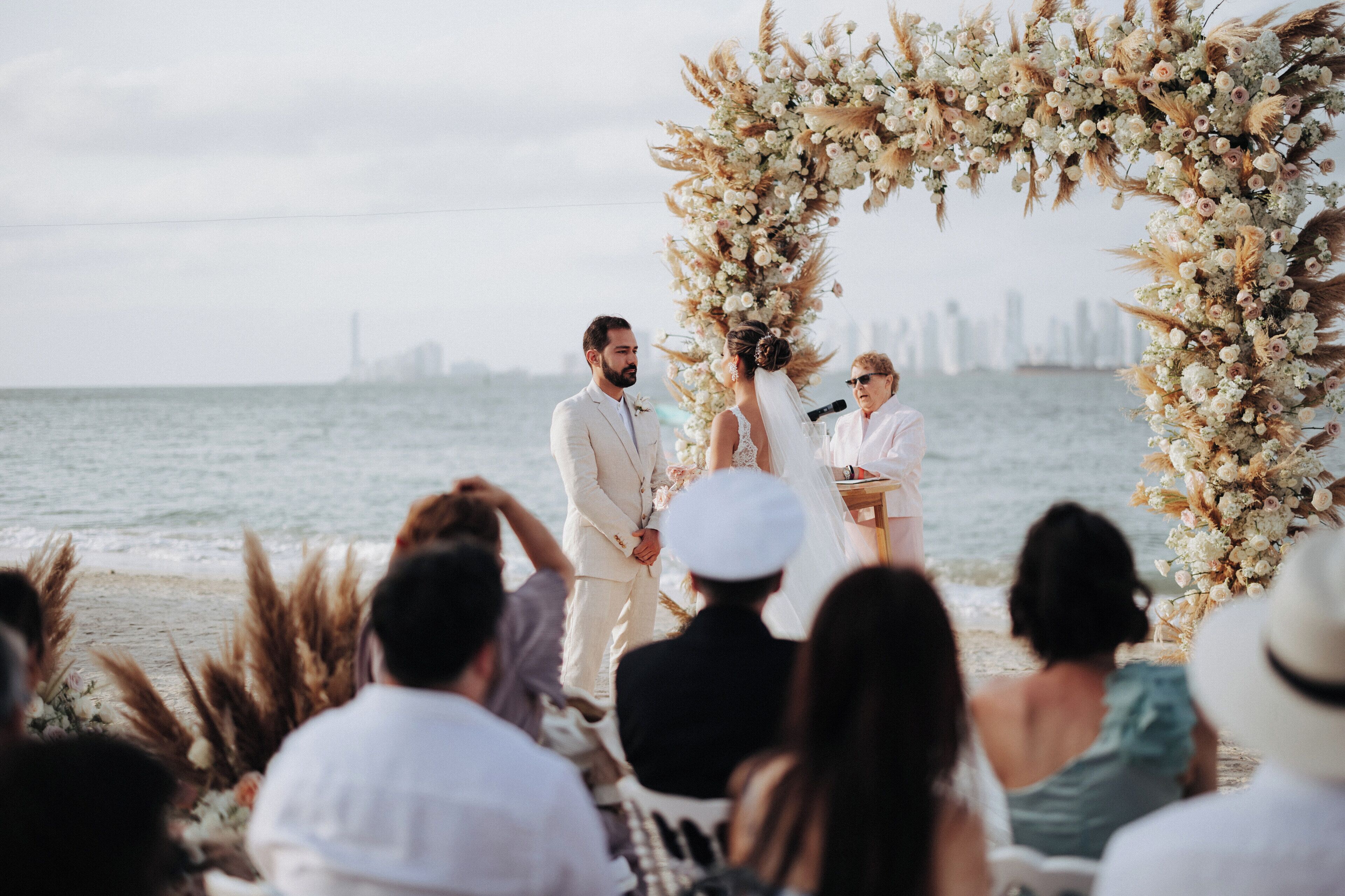 Une cérémonie de mariage sur une plage, avec une arche fleurie et la silhouette de la ville en arrière-plan.