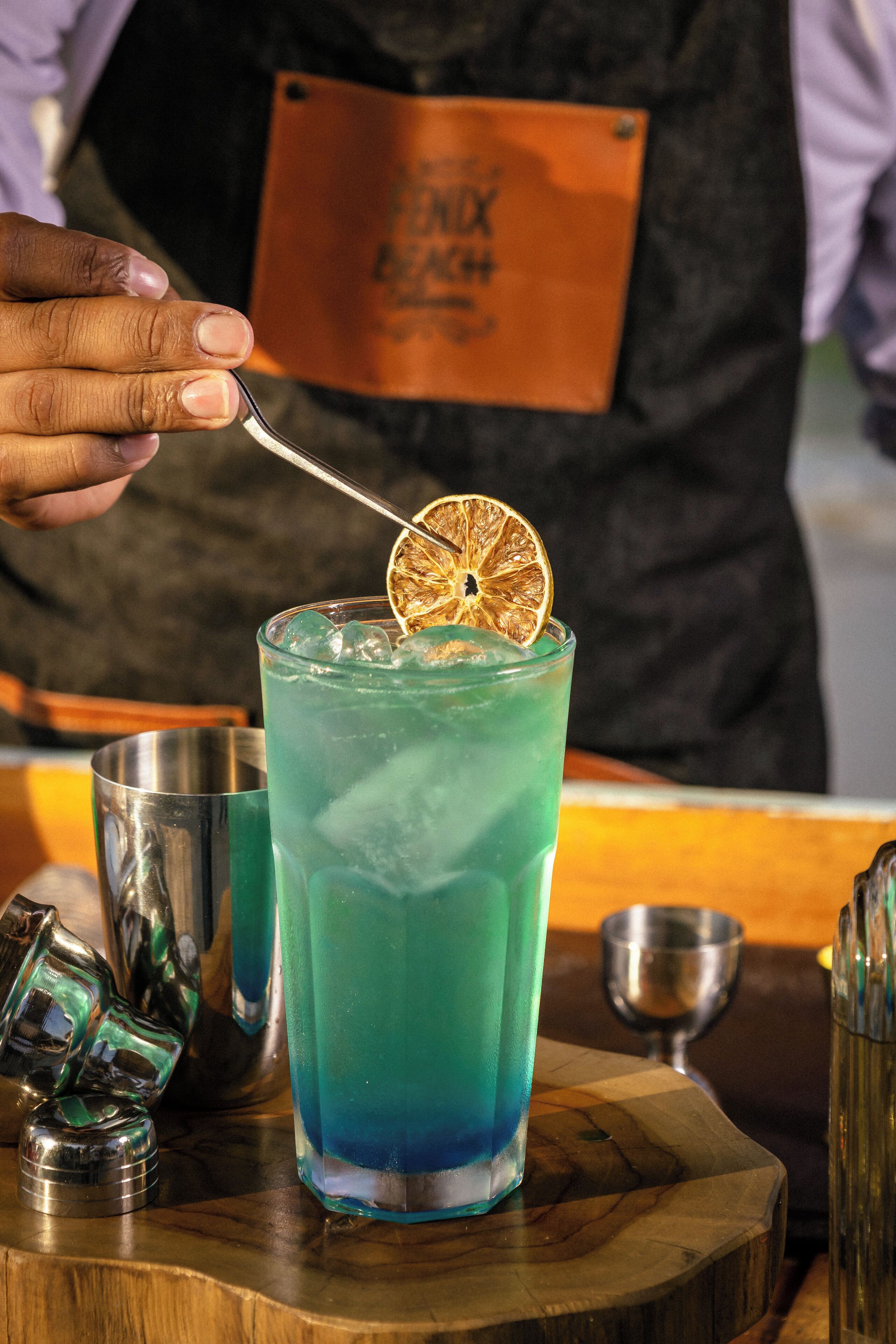 A bartender preparing a blue cocktail with a garnish, surrounded by bar tools on a wooden surface.