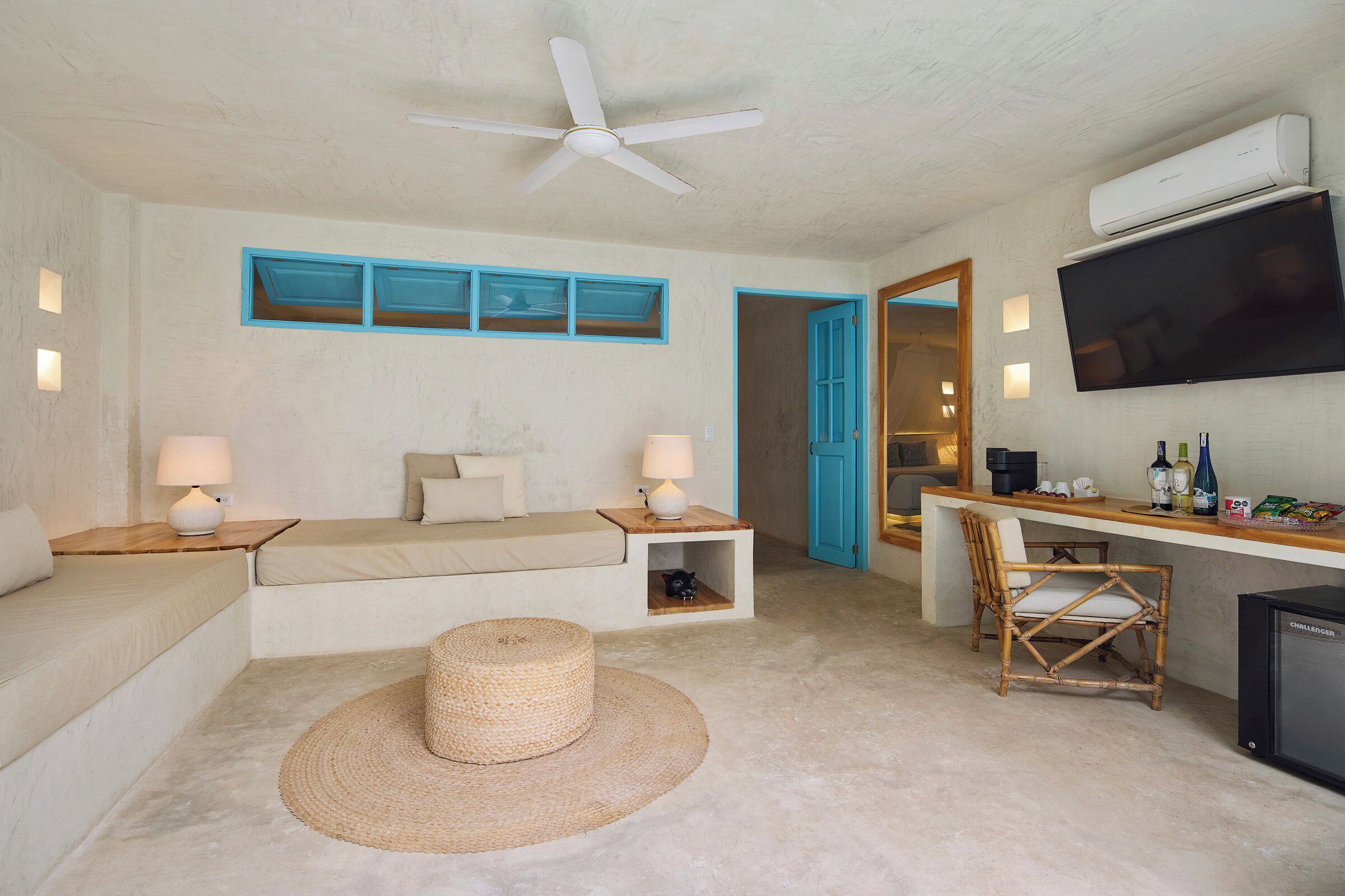 A room with a sofa, a small table with a lamp, a wicker ottoman, a TV, a desk with a chair, and an air conditioning unit.