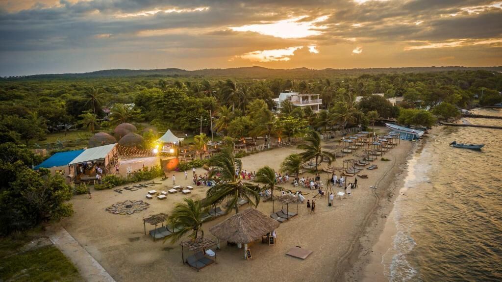 Un resort frente a la playa con varias cabañas, un espacio central abierto con una tienda de campaña y una playa con sombrillas y sillas de descanso.