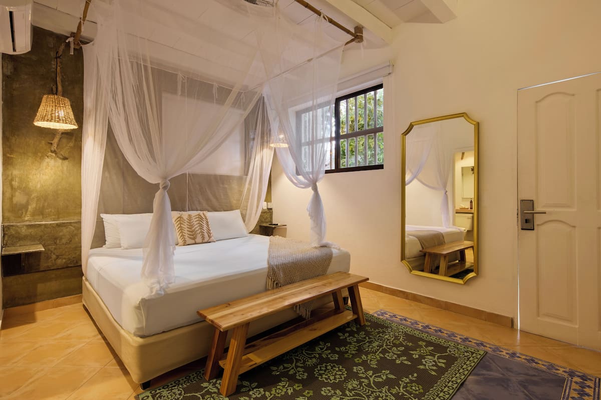 Une chambre à coucher comprenant un lit, une table de chevet, un banc, un baldaquin, une fenêtre, un miroir et une porte.