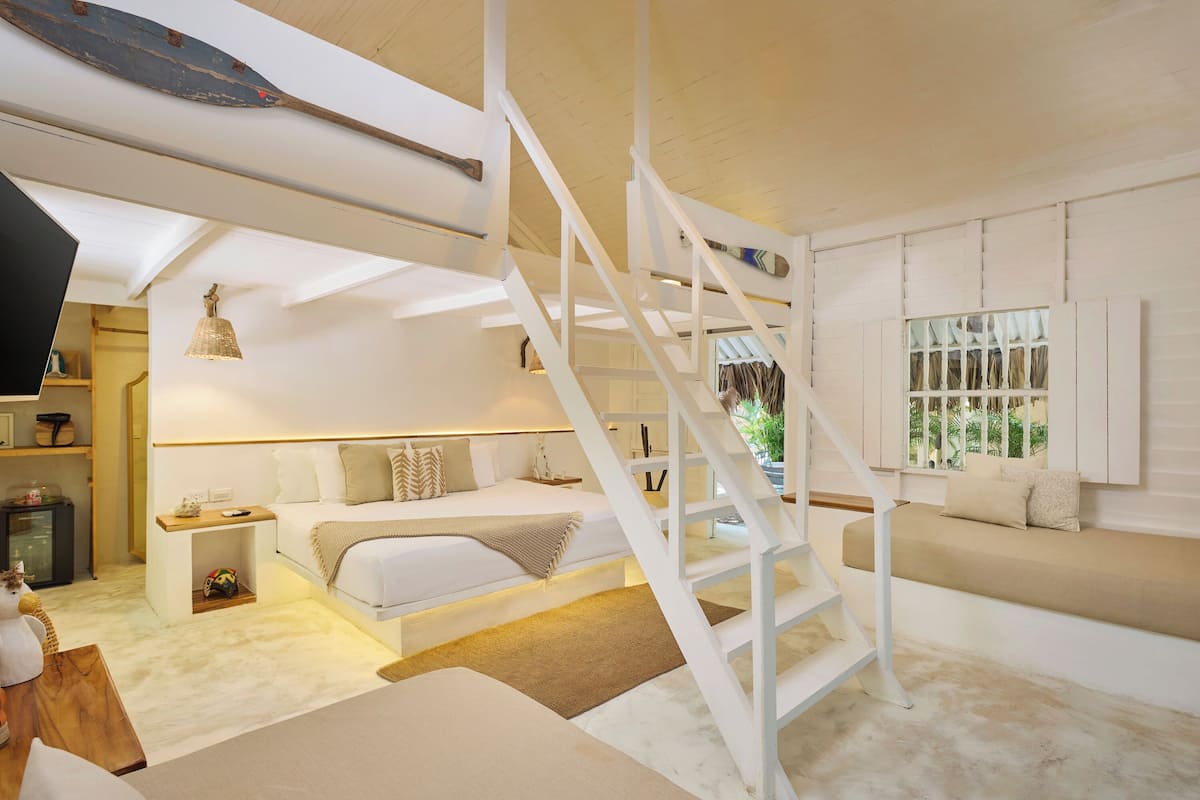 Une chambre à coucher avec un lit, des tables de chevet, un banc en bois, un escalier et une fenêtre avec des volets.