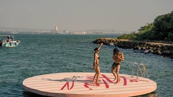 Eine runde, schwimmende Plattform mit Geländer und einer Leiter, in einem Gewässer mit einer entfernten Skyline im Hintergrund.
