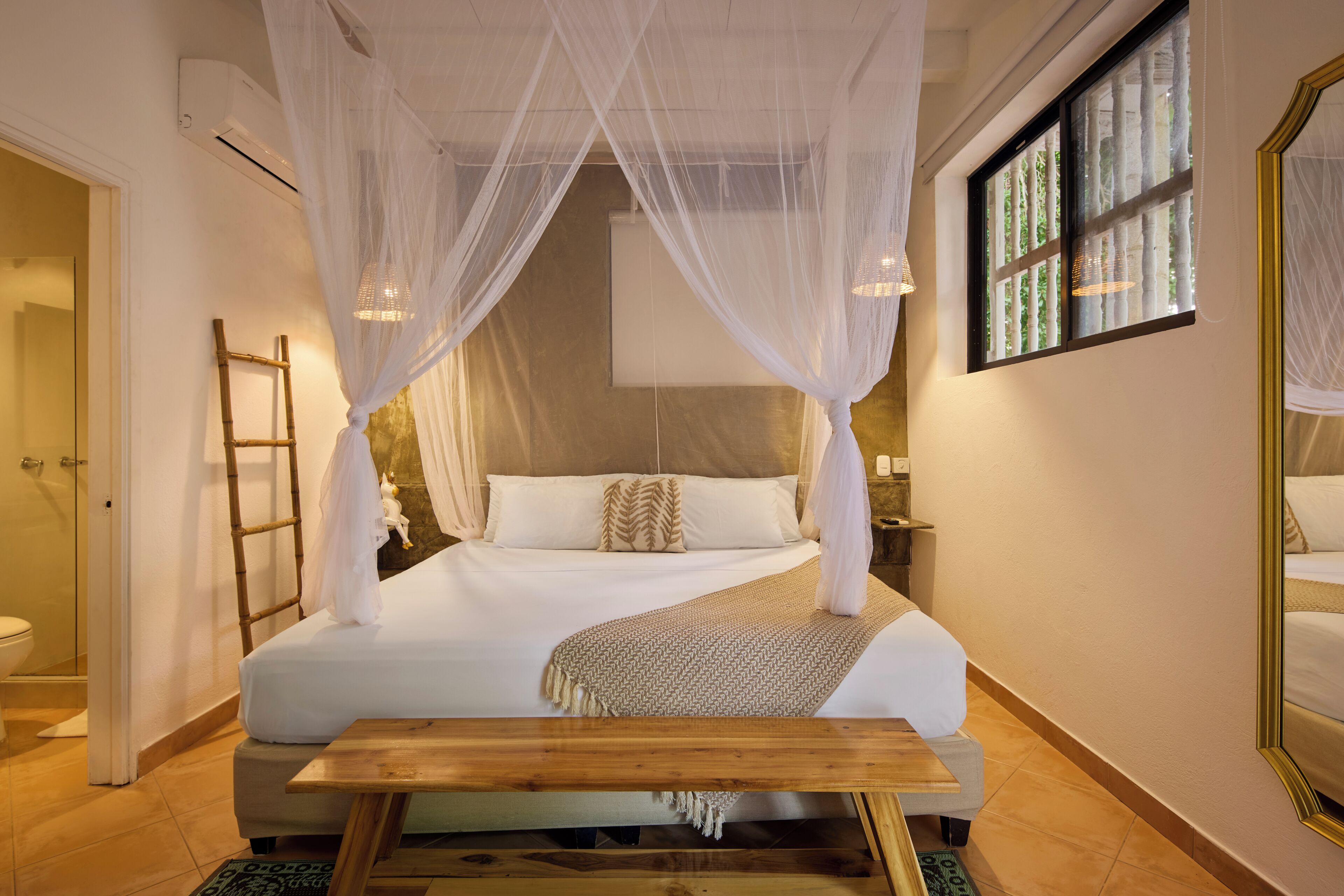 A bedroom with a four-poster canopy bed, a wooden bench, a bedside table, and a large mirror.