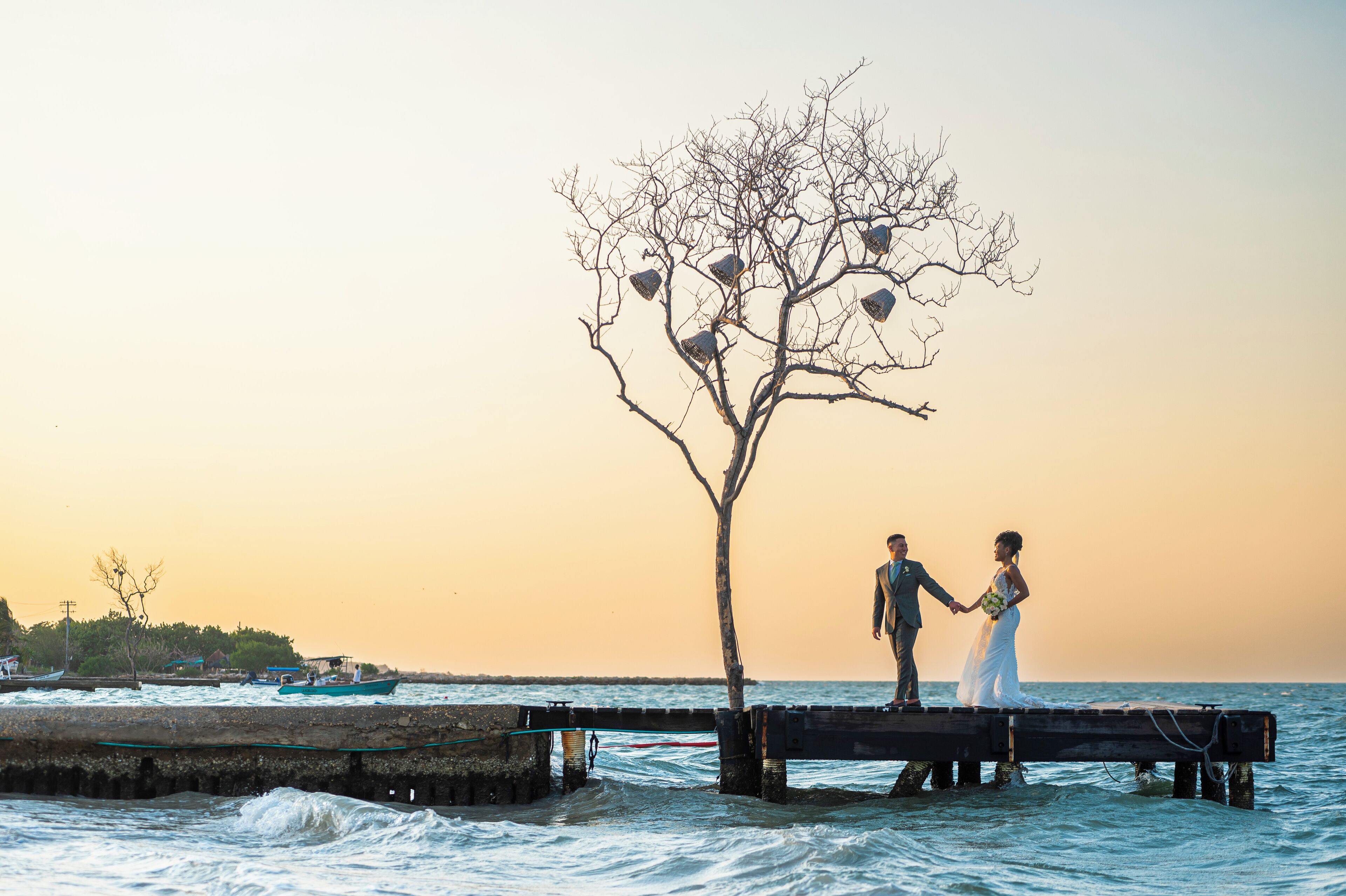 A couple holding hands on a wooden pier by the sea with boats in the background.