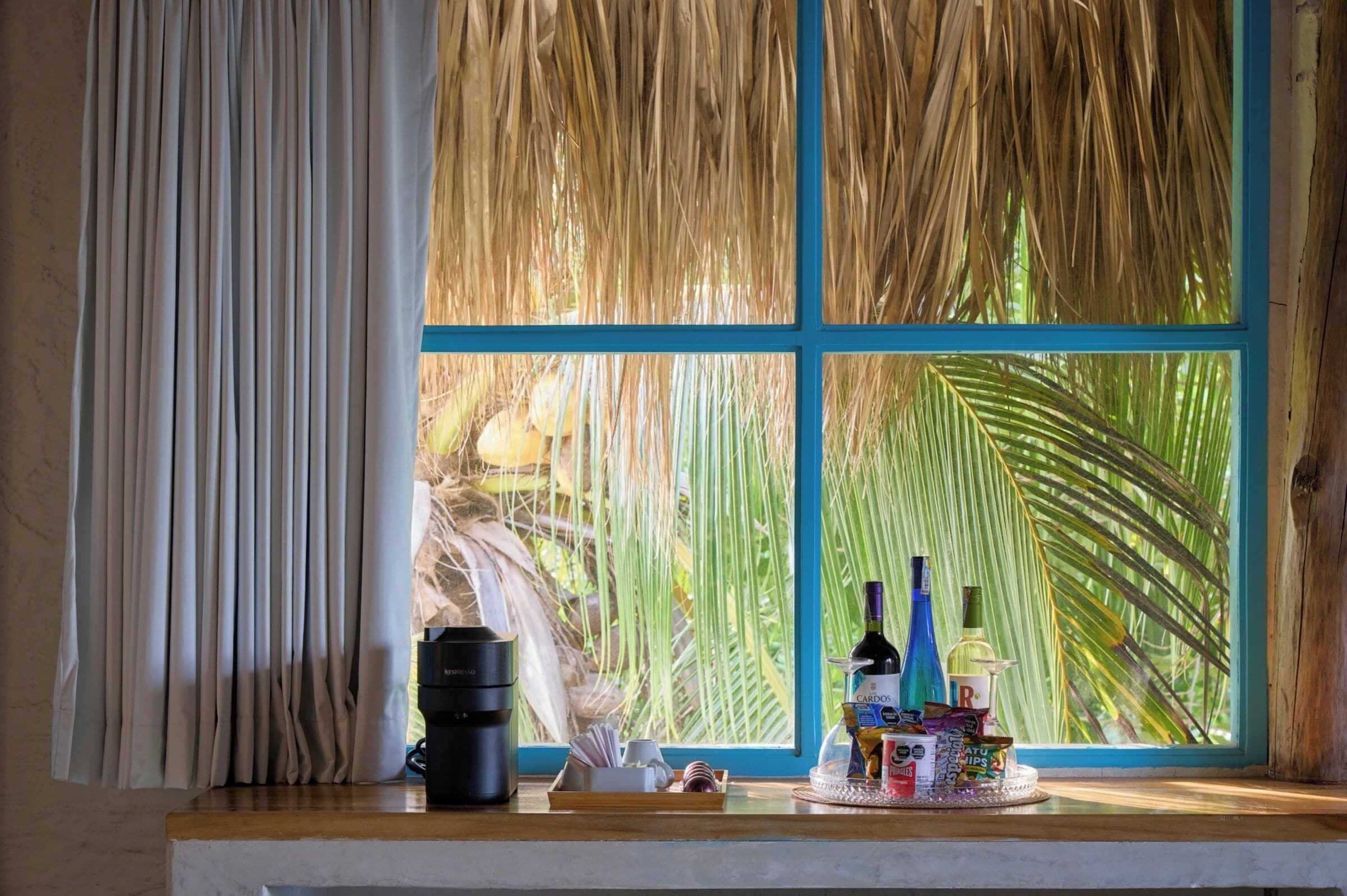 A window with a view of palm trees, a coffee maker, and a tray with bottles and snacks.