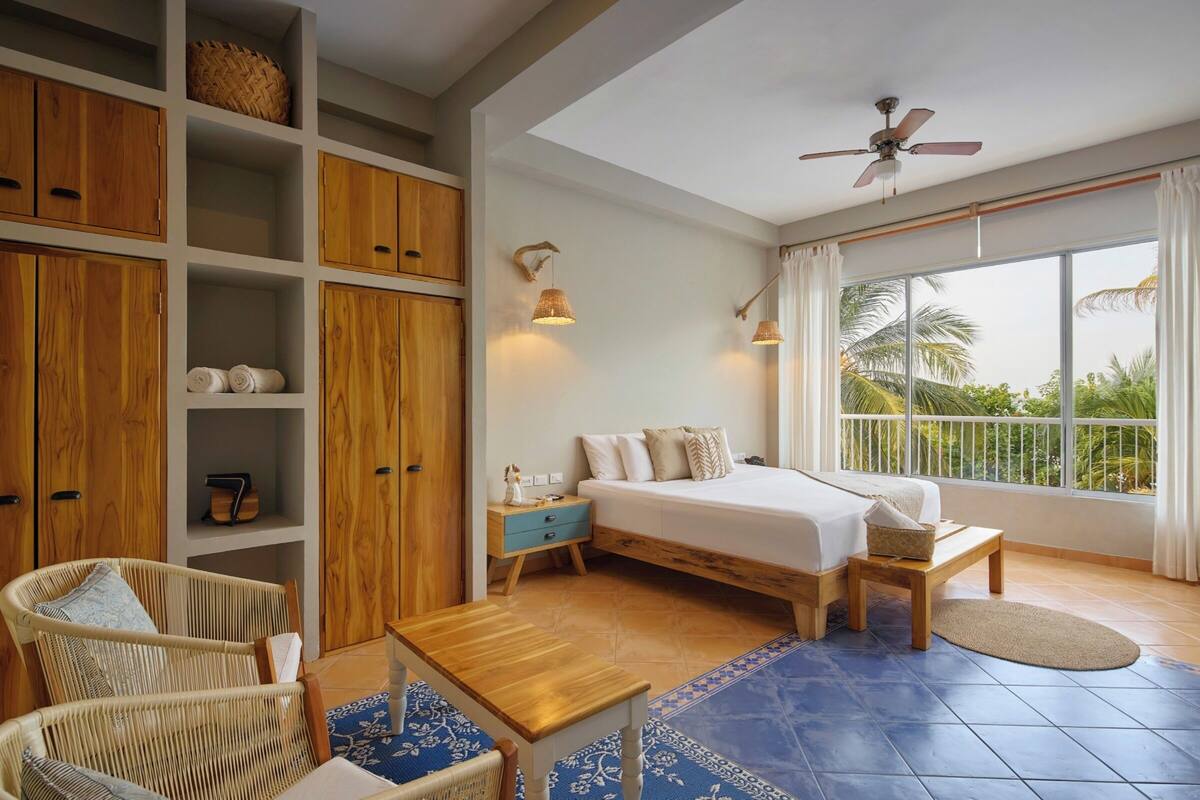 Une chambre spacieuse avec un grand lit, une armoire en bois, des fauteuils en osier, une petite table et une vue sur les palmiers à travers de grandes fenêtres.