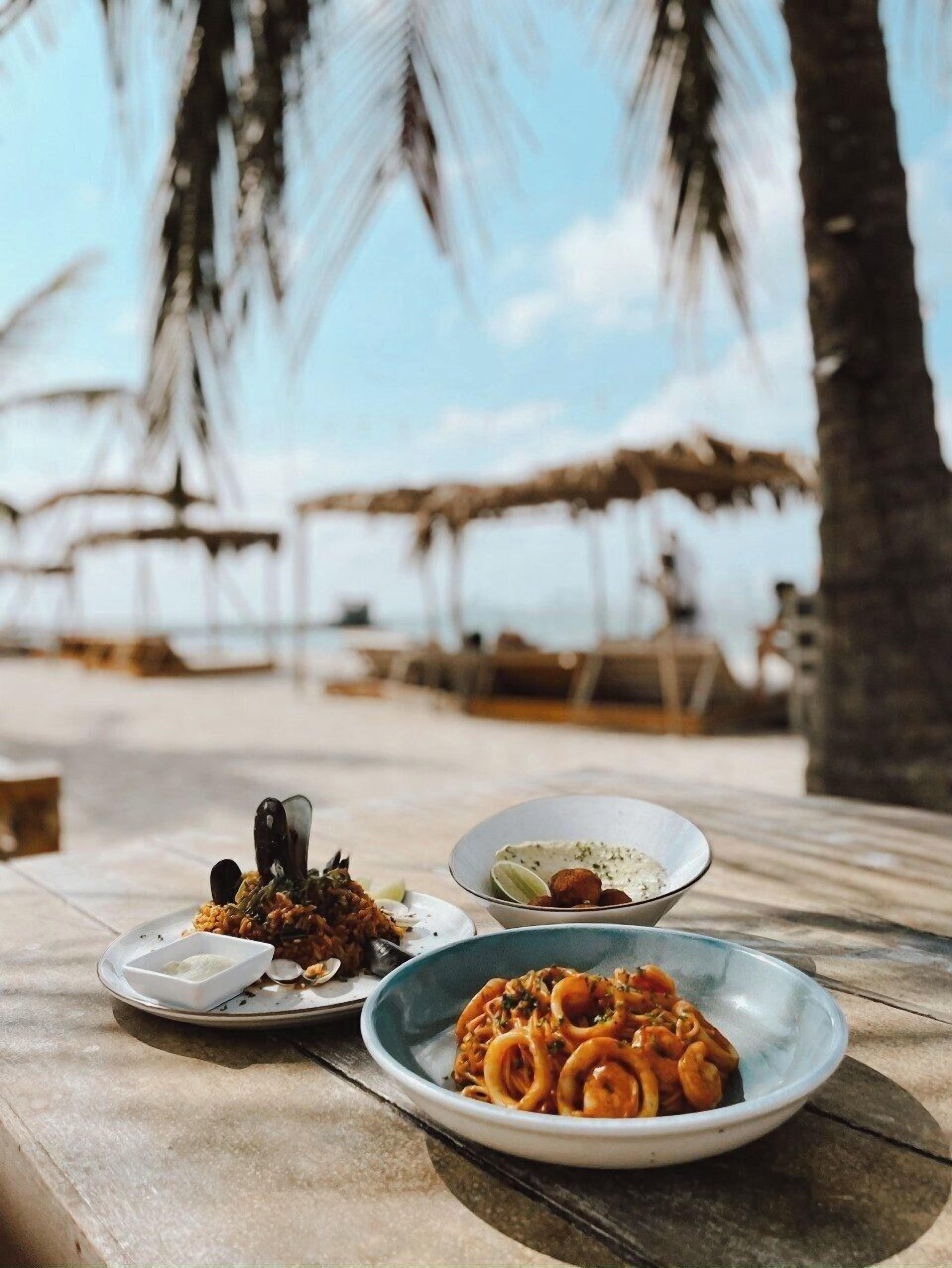 A table with three plates of food, a bowl of pasta, and a small dish of rice, set outdoors with palm trees in the background.