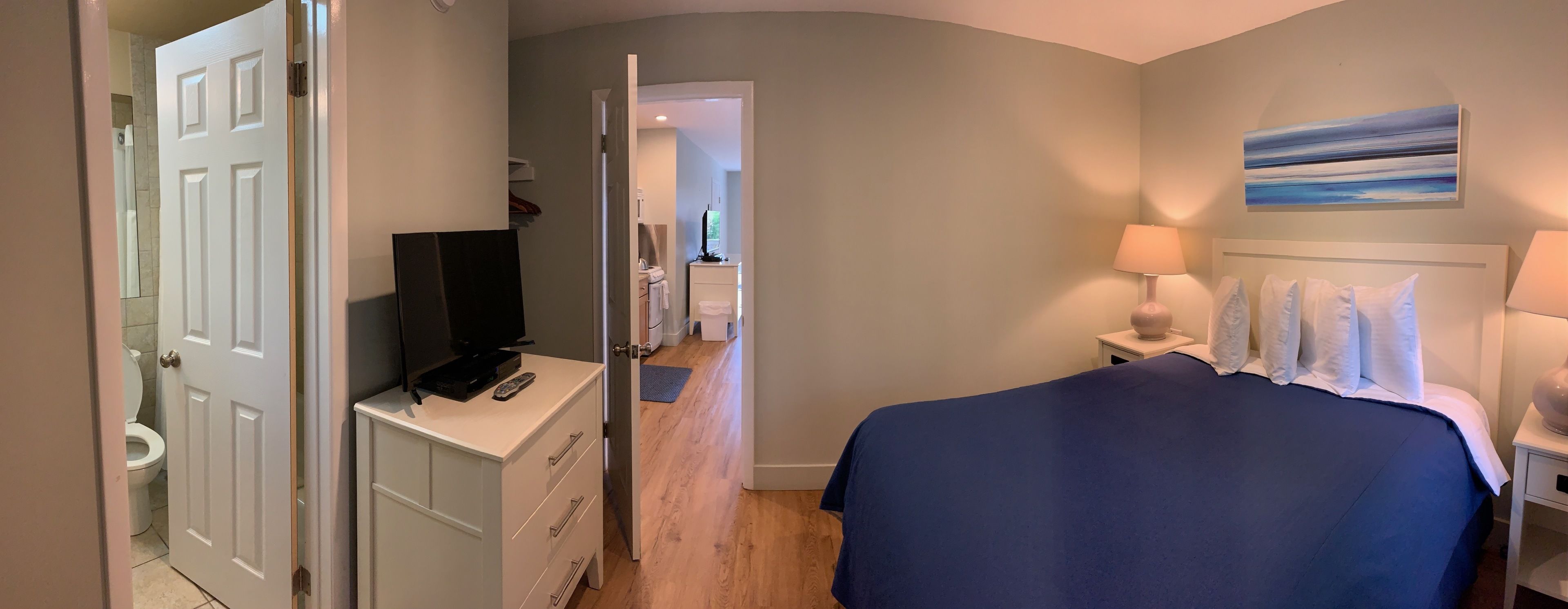 Ett sovrum med en säng, en tv-apparat på en byrå, ett badrum som syns genom en öppen dörr och en tavla på väggen.
