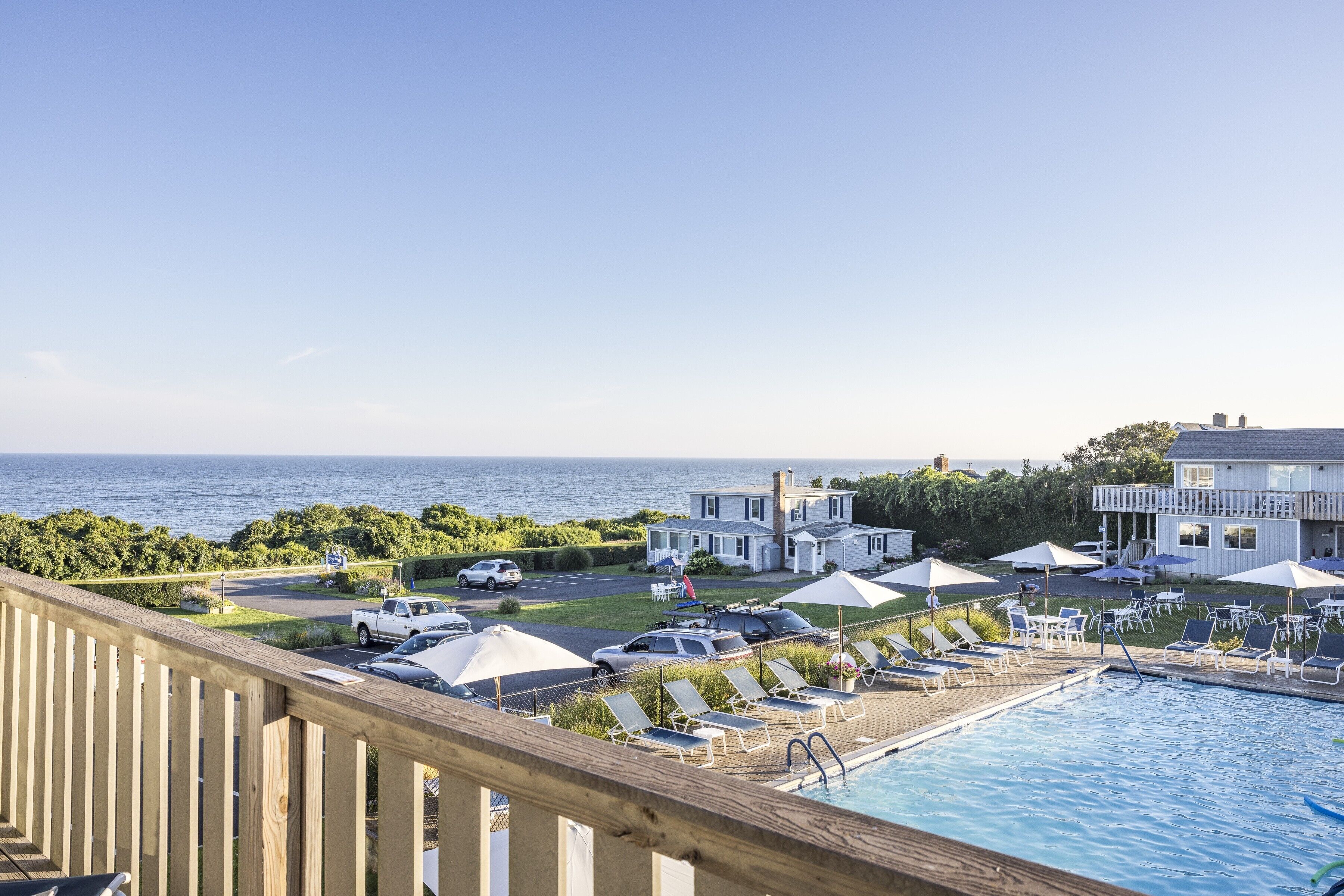 A poolside area with lounge chairs and umbrellas, a view of the sea, and a parking lot with vehicles.