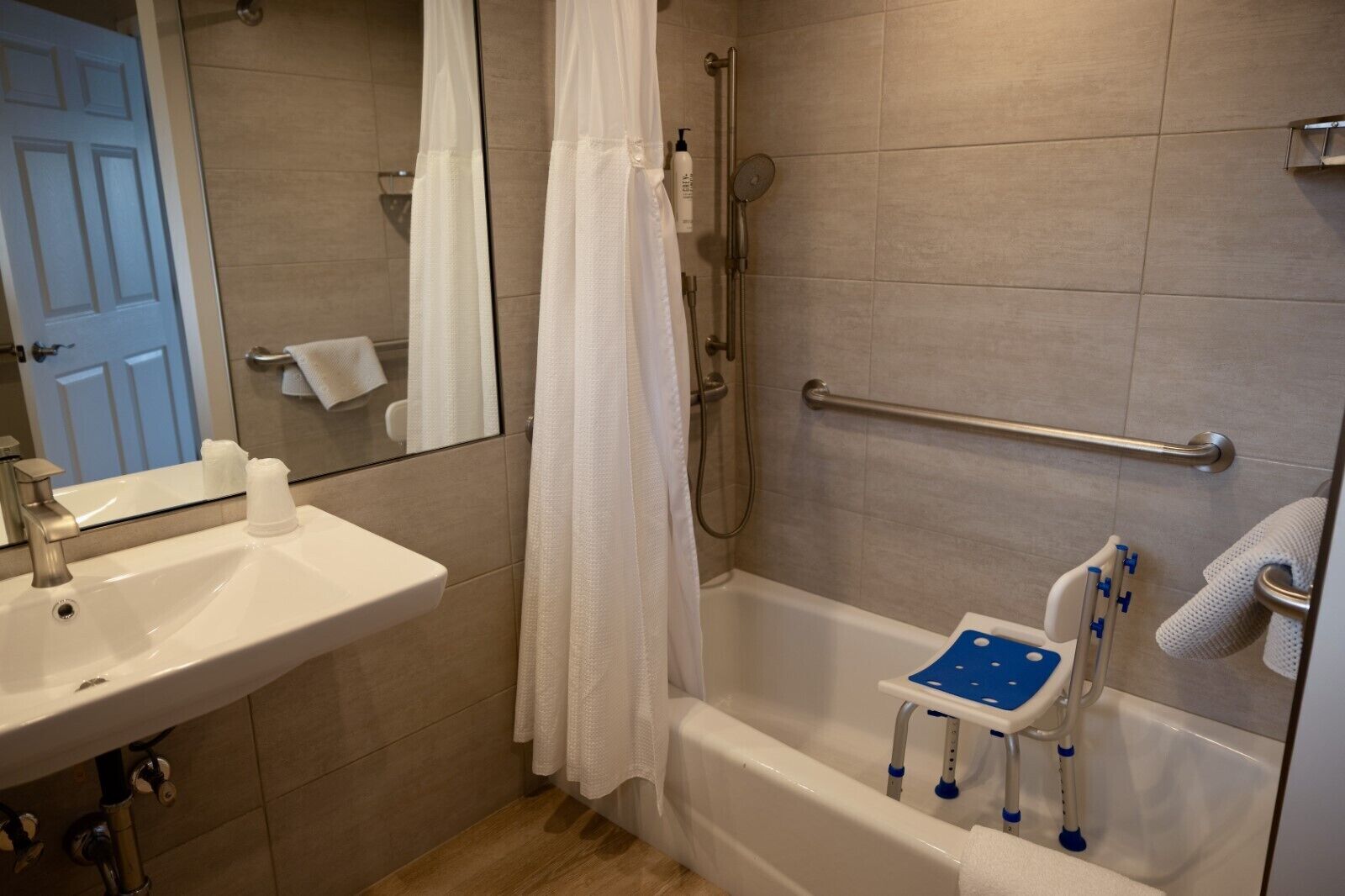 A bathroom with a white sink, a shower with a handheld showerhead, a bathtub with a blue seat, and a white towel rack.