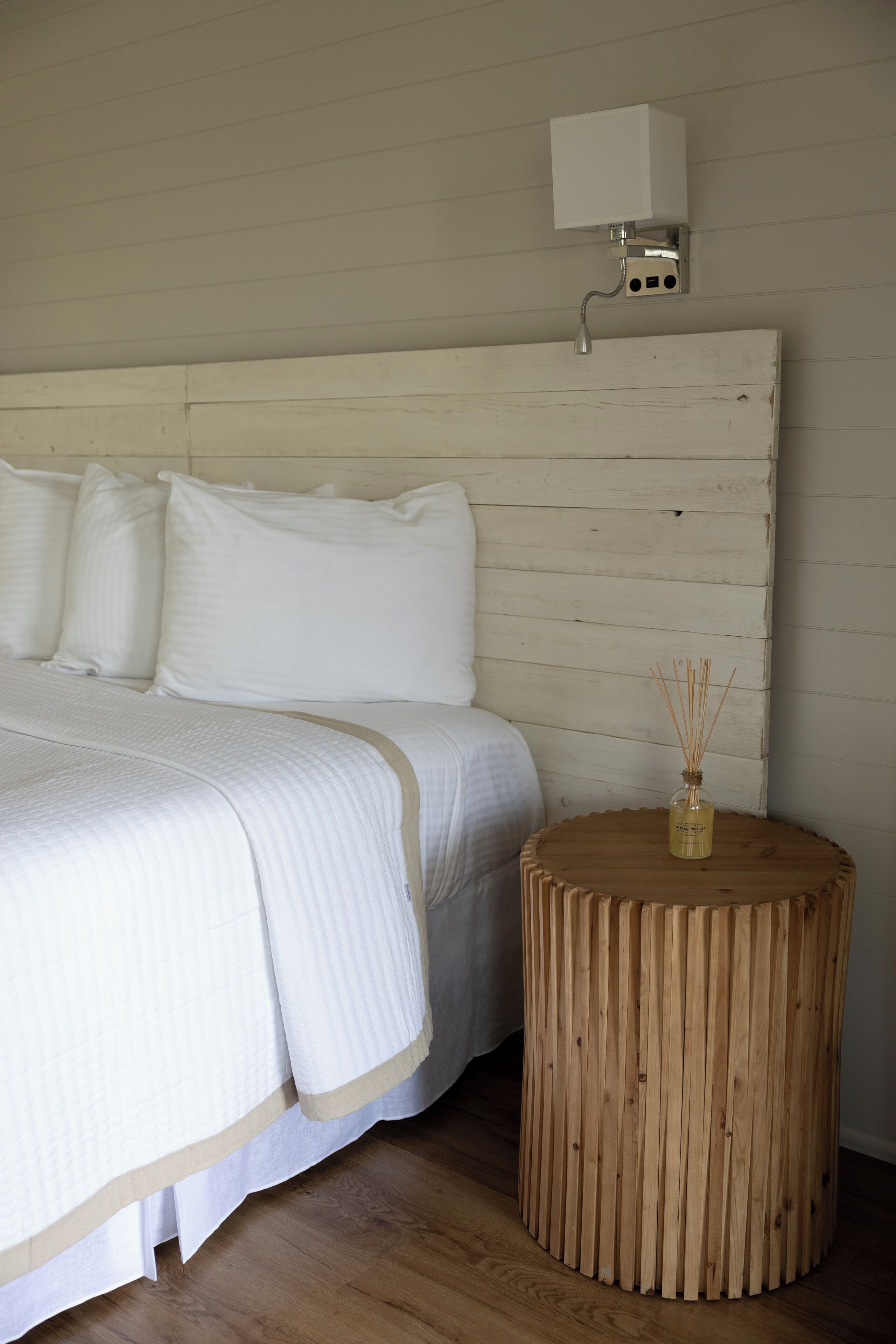 Un dormitorio con cabecera de madera, una mesita de noche redonda de madera y una colcha blanca.