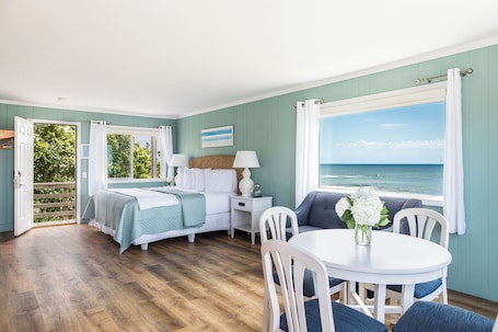 A room with a bed, a table with chairs, a window with a view of the ocean, and a door leading to an outdoor area.