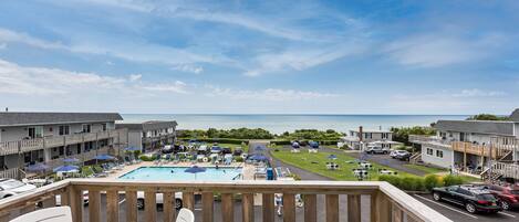 A pool area with lounge chairs and umbrellas, a view of the sea, and a parking lot with cars.