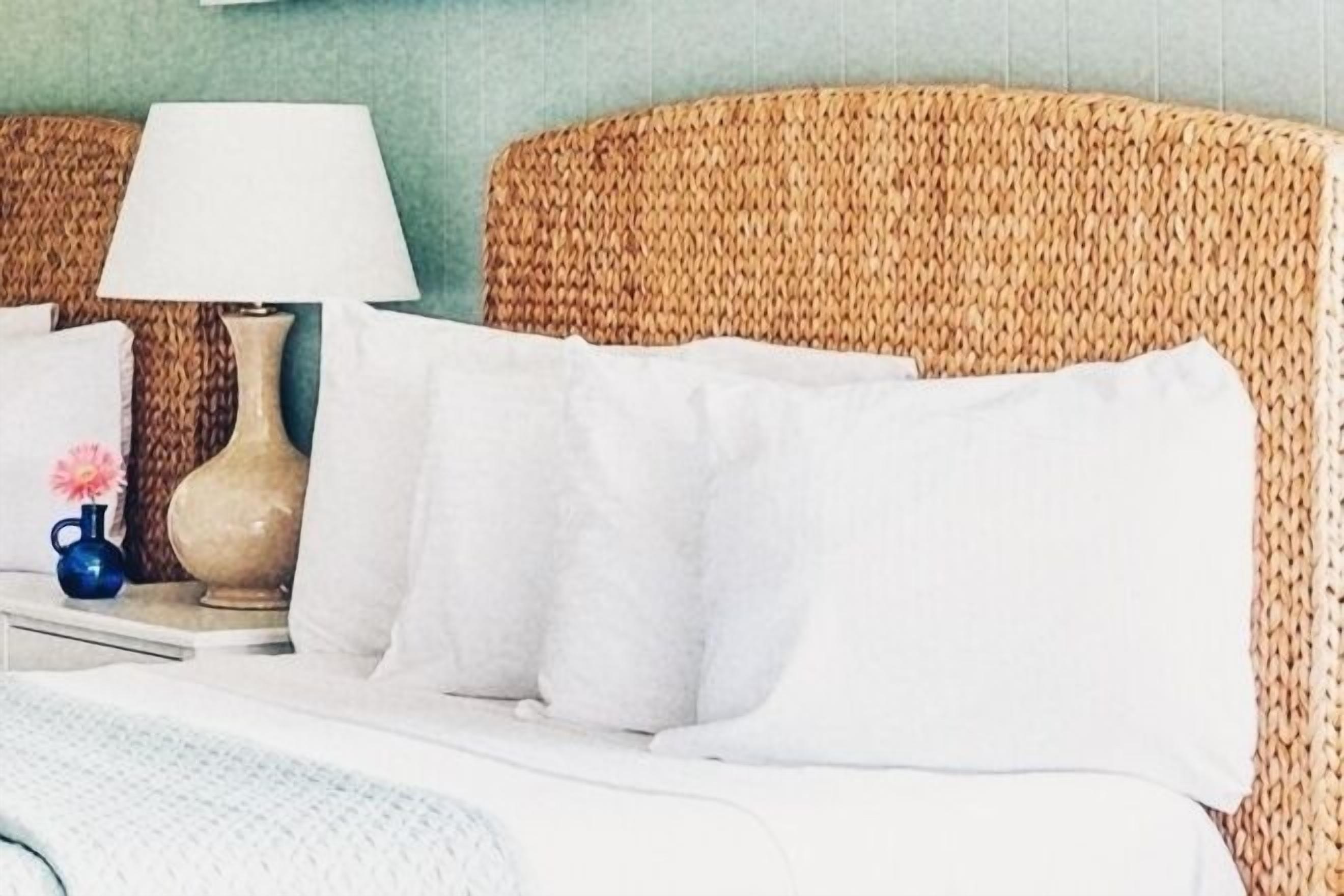 Una cama bien hecha con sábanas y almohadas blancas, una lámpara de noche y un florero con una flor rosa sobre una mesita de noche.