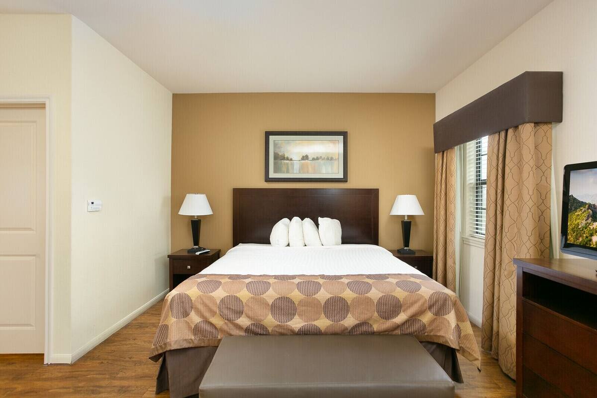 A hotel room with a bed, two bedside tables with lamps, a bench, a painting on the wall, and a television on a stand.