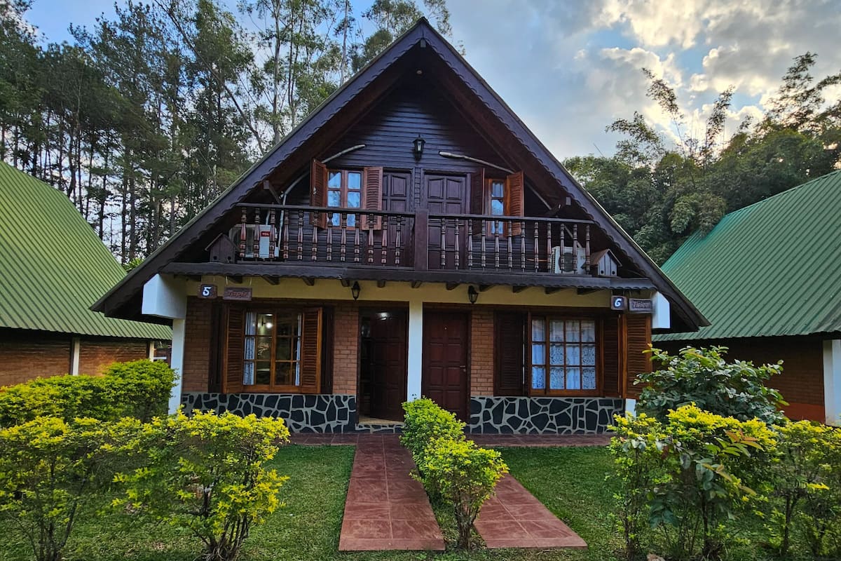A two-story wooden house with a balcony, surrounded by greenery and a stone pathway leading to the entrance.