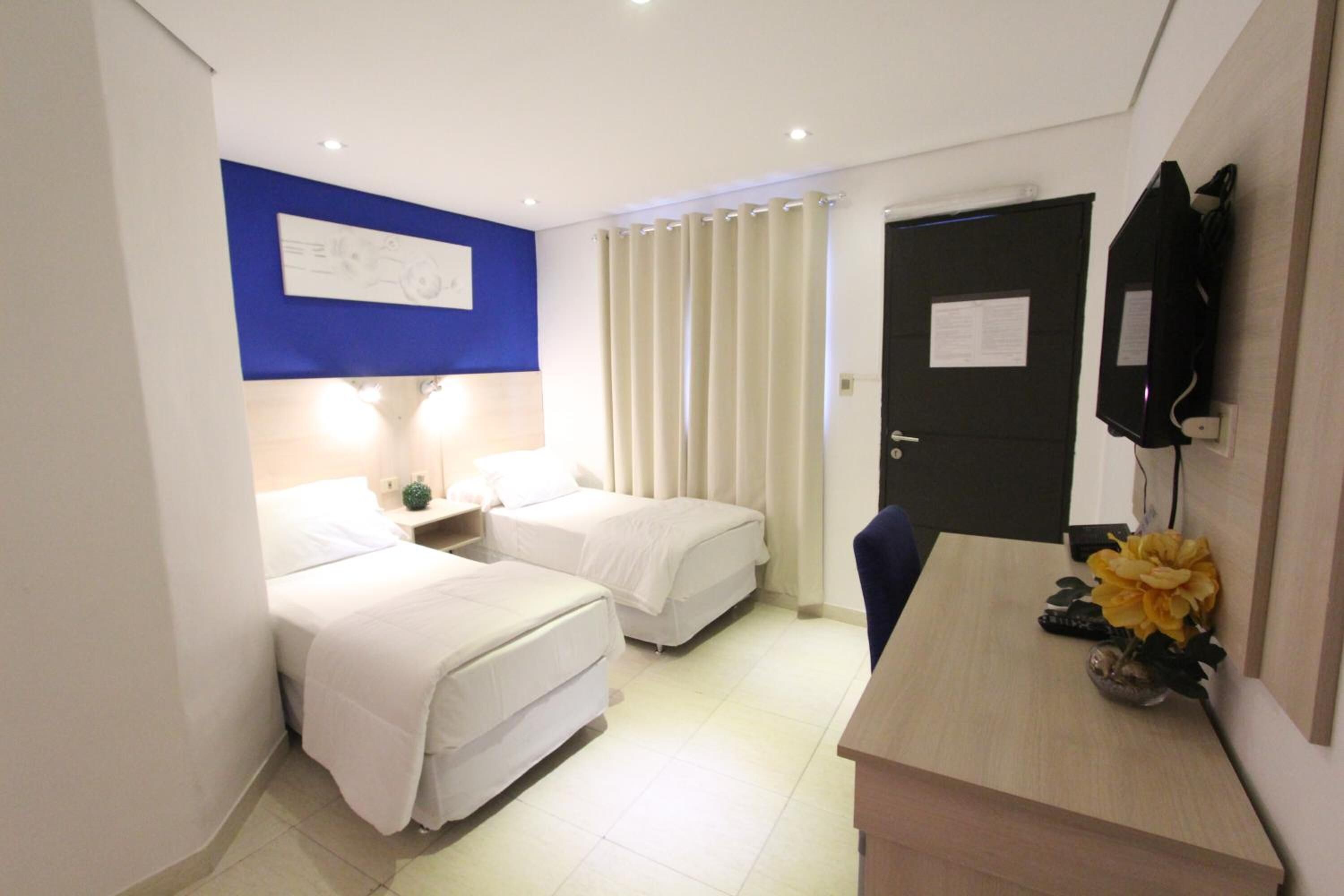 A hotel room with two beds, a desk, a TV, and a door with a notice.