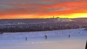 A snowy outdoor area with a metal railing and a wooden deck leading to a view of a sunset over a town. - Cozy Nest B&B (Calgary)