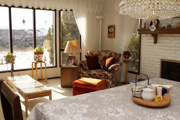 A cozy living room with a fireplace, a dining table, and a view of a snowy landscape through large windows.
