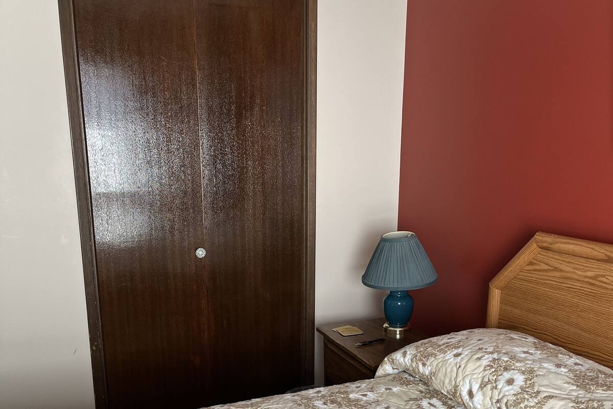A bedroom with a wooden wardrobe, a bedside table with a lamp, and a floral-patterned bedspread.