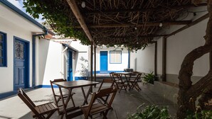 Un espacio exterior con mesas y sillas de madera, rodeado de paredes blancas con detalles azules y puertas.