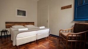 Un dormitorio ordenado con suelo de madera, una cama con sábanas blancas, una silla de madera y un cuadro en la pared.