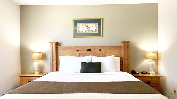 A neatly made bed with a wooden headboard, two bedside lamps, a nightstand with a phone and a clock, and a framed picture on the wall. - Burke Inn Motel & Suites (Carroll)