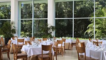 A restaurant with multiple tables set for dining, large windows offering a view of lush greenery.